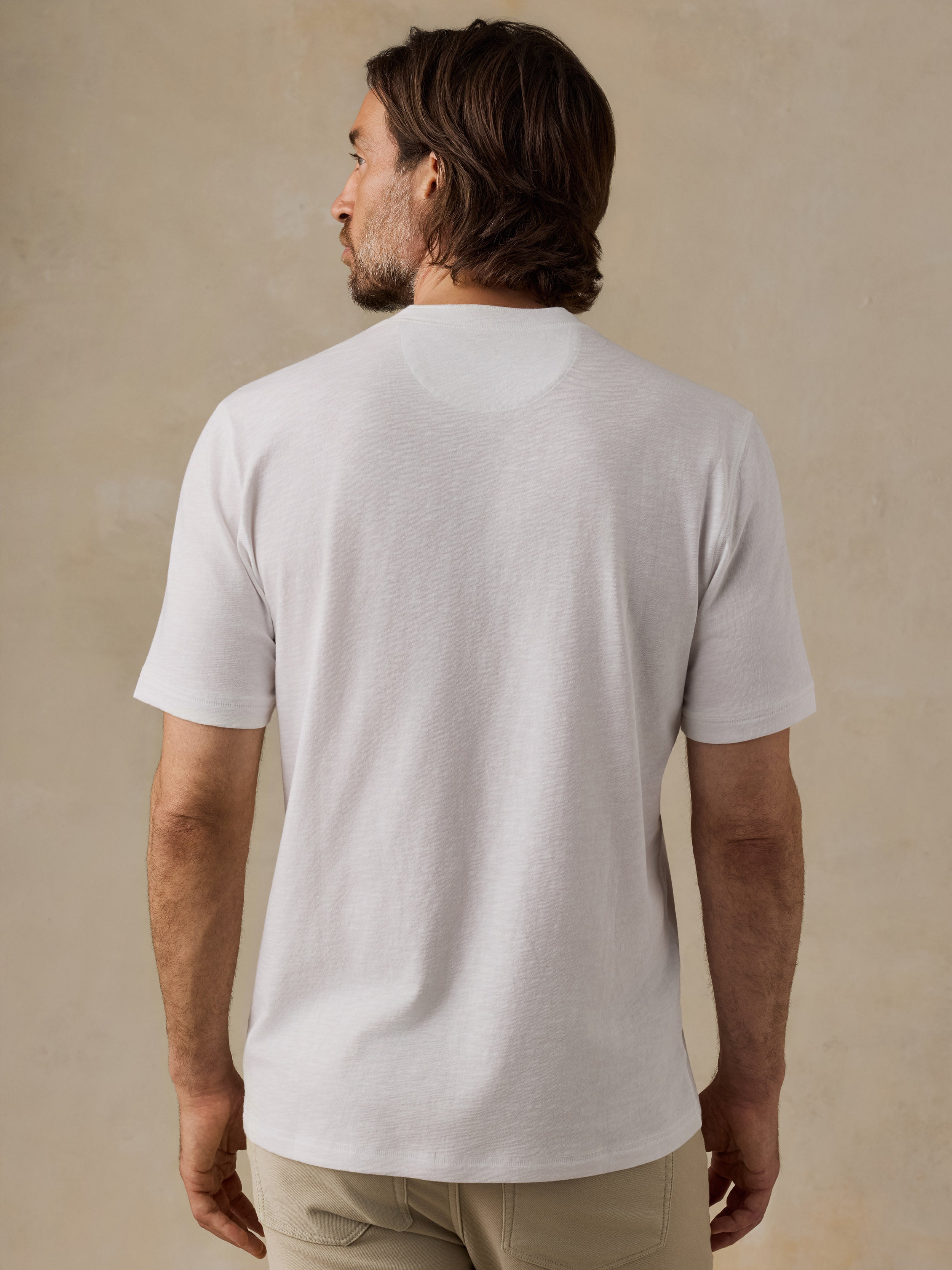 A man viewed from the back wearing a plain white short-sleeve crewneck t-shirt and khaki pants, standing against a textured beige studio wall.