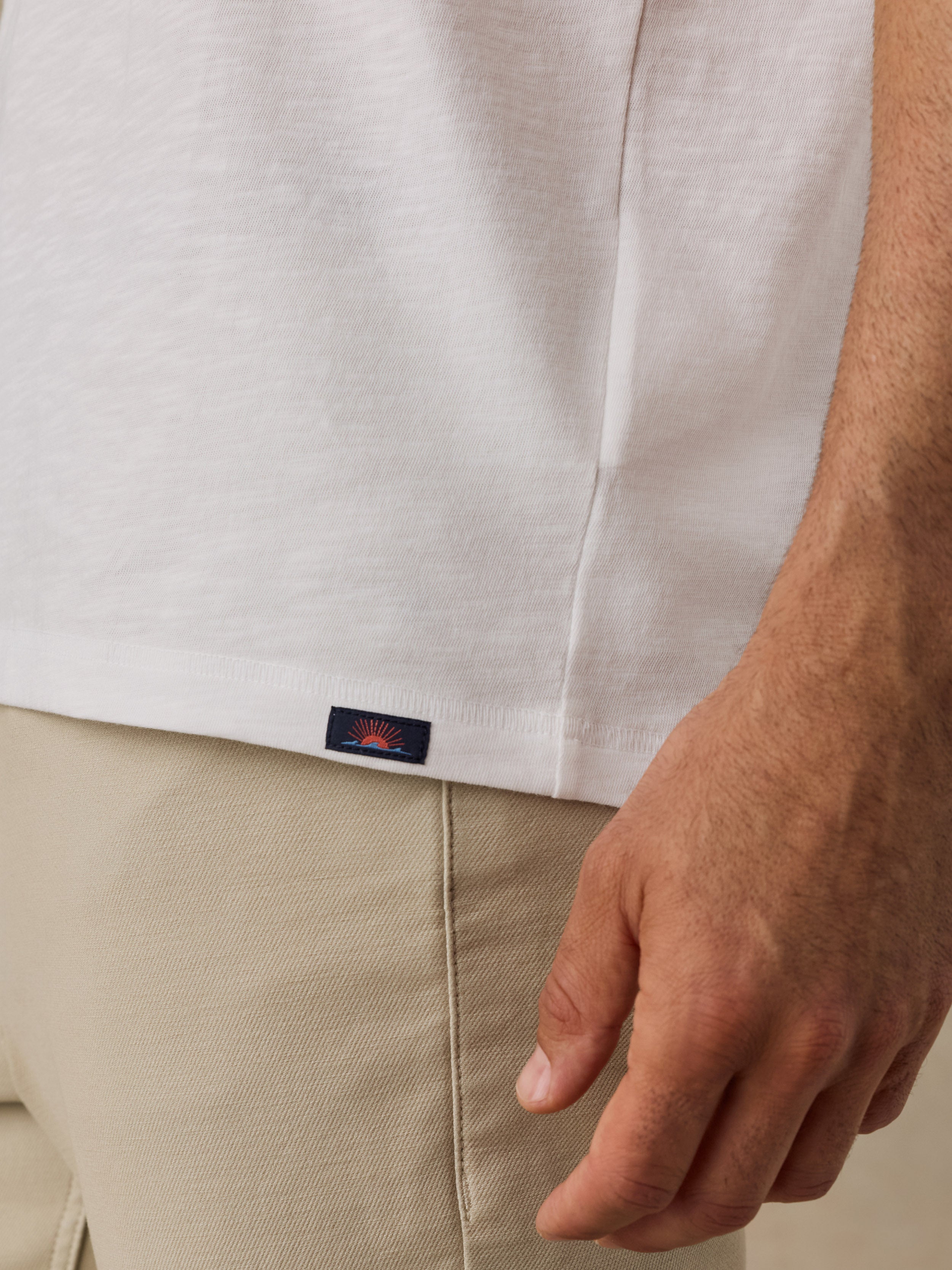 Close-up of a man wearing a white short-sleeve t-shirt and beige trousers, with a small navy Faherty brand patch on the hem of the shirt.