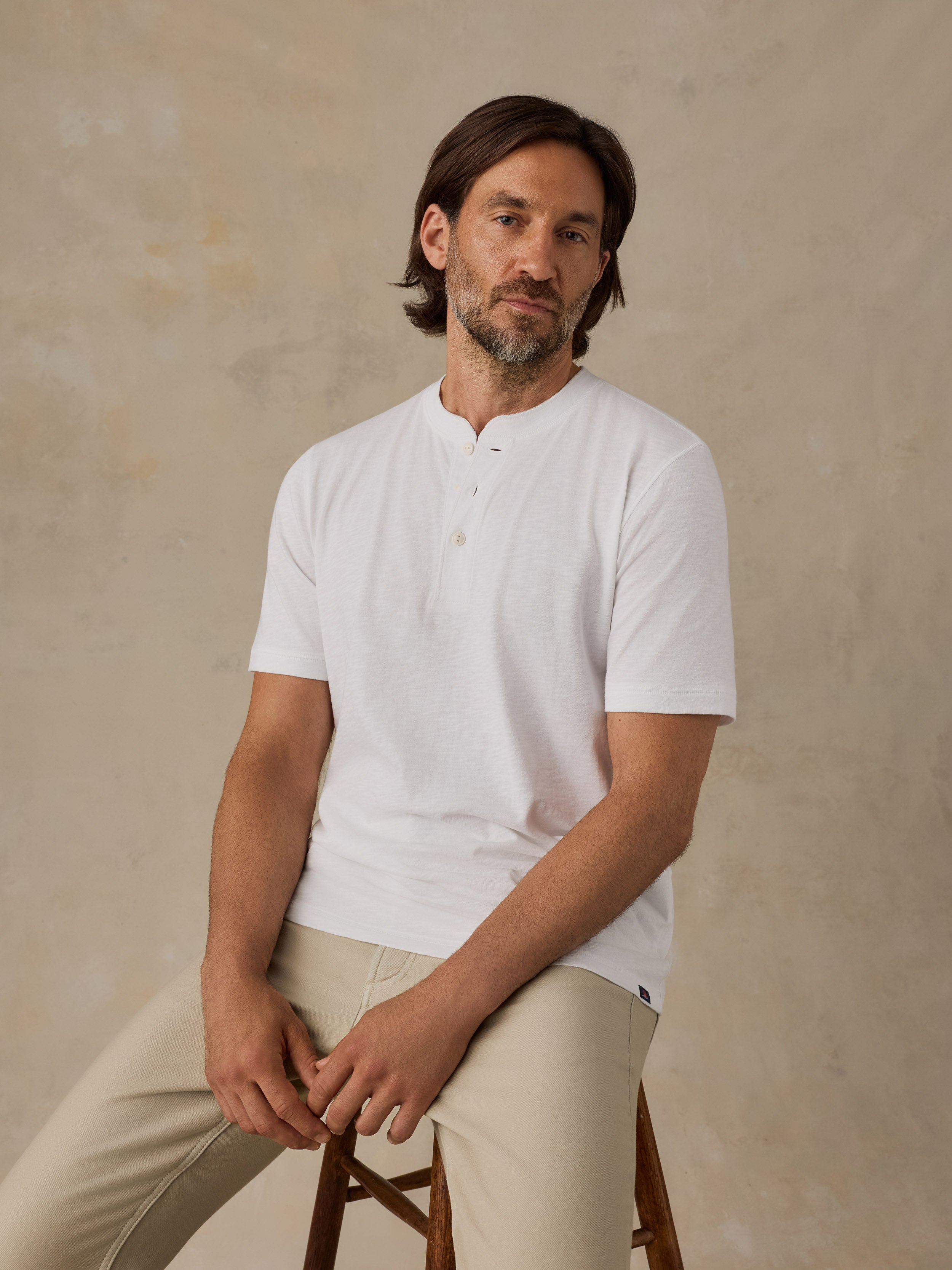 A man wearing a white short-sleeve henley shirt and beige trousers, sitting on a wooden stool against a textured beige studio backdrop.