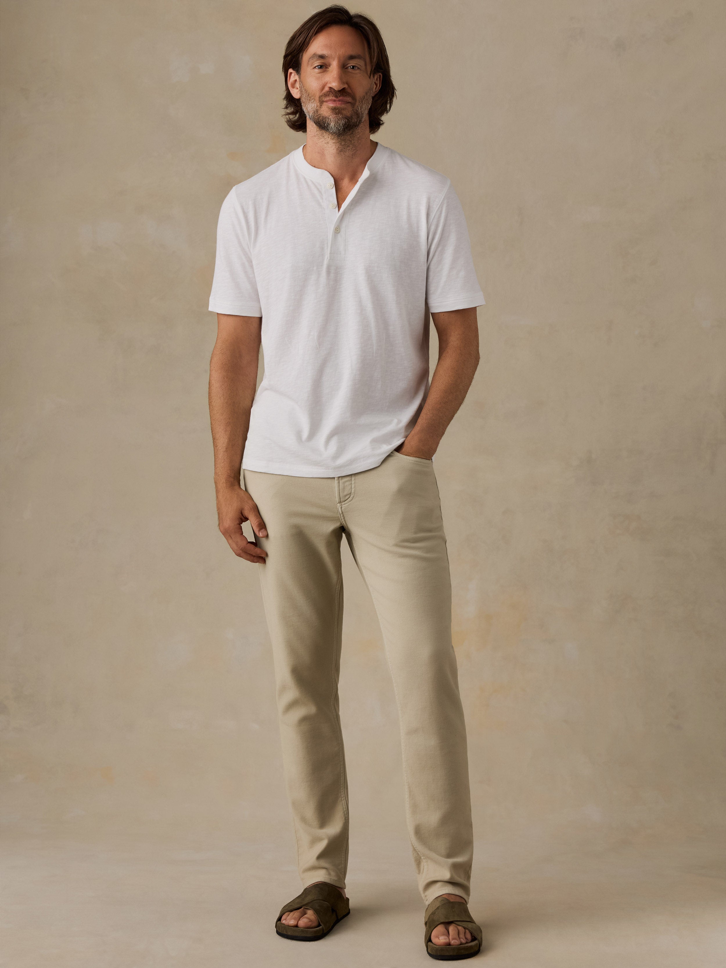 A man wearing a white short-sleeve henley shirt, beige trousers, and brown sandals, standing against a textured beige studio backdrop.