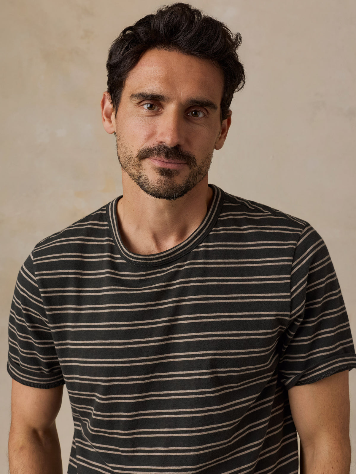 A man wearing a short-sleeve crewneck t-shirt with thin horizontal stripes in black and beige, set against a neutral textured beige background., image 1 of 6