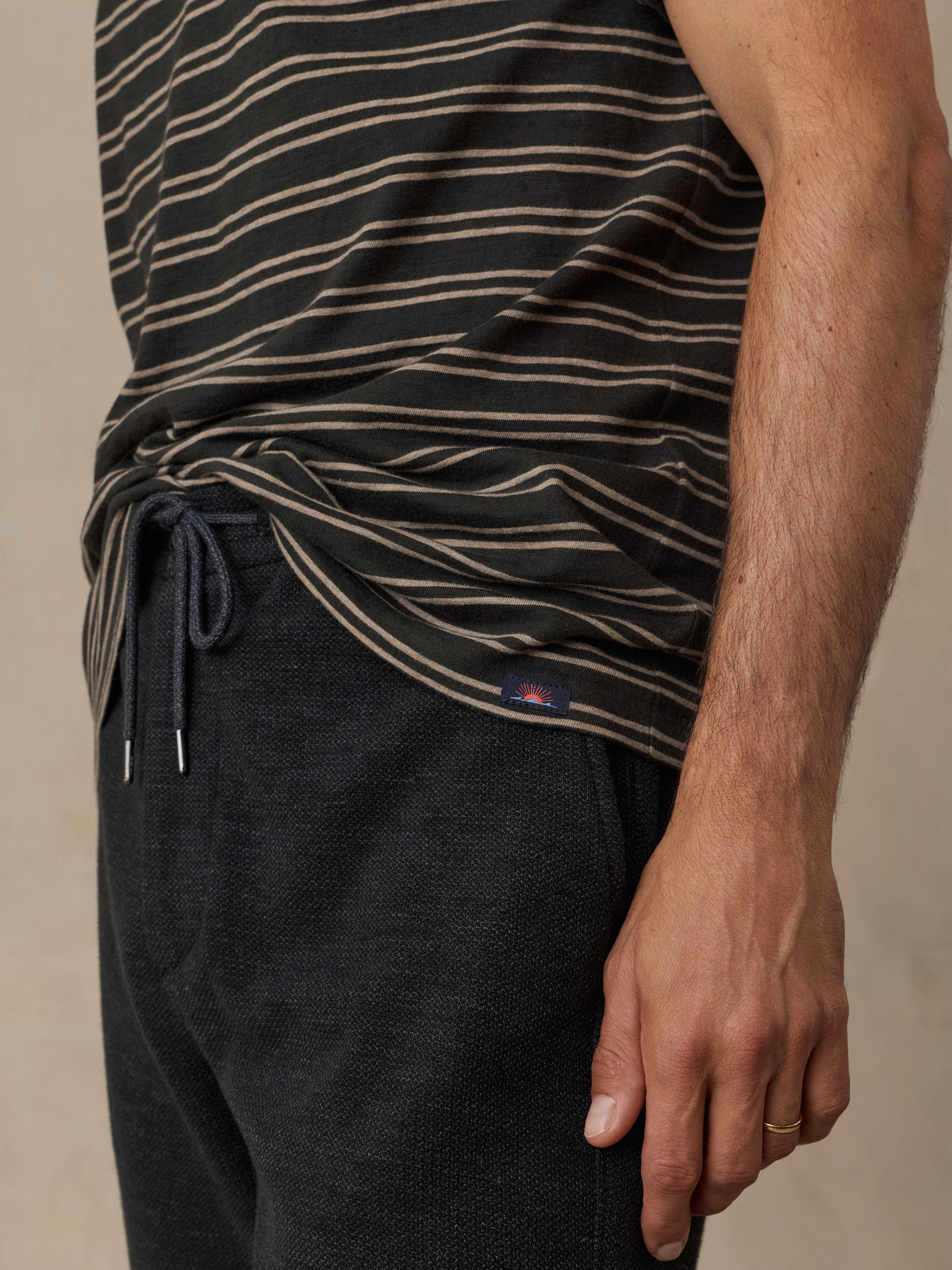 Close-up of a man wearing a black striped t-shirt and dark charcoal knit pants against a beige studio background, image 4 of 6
