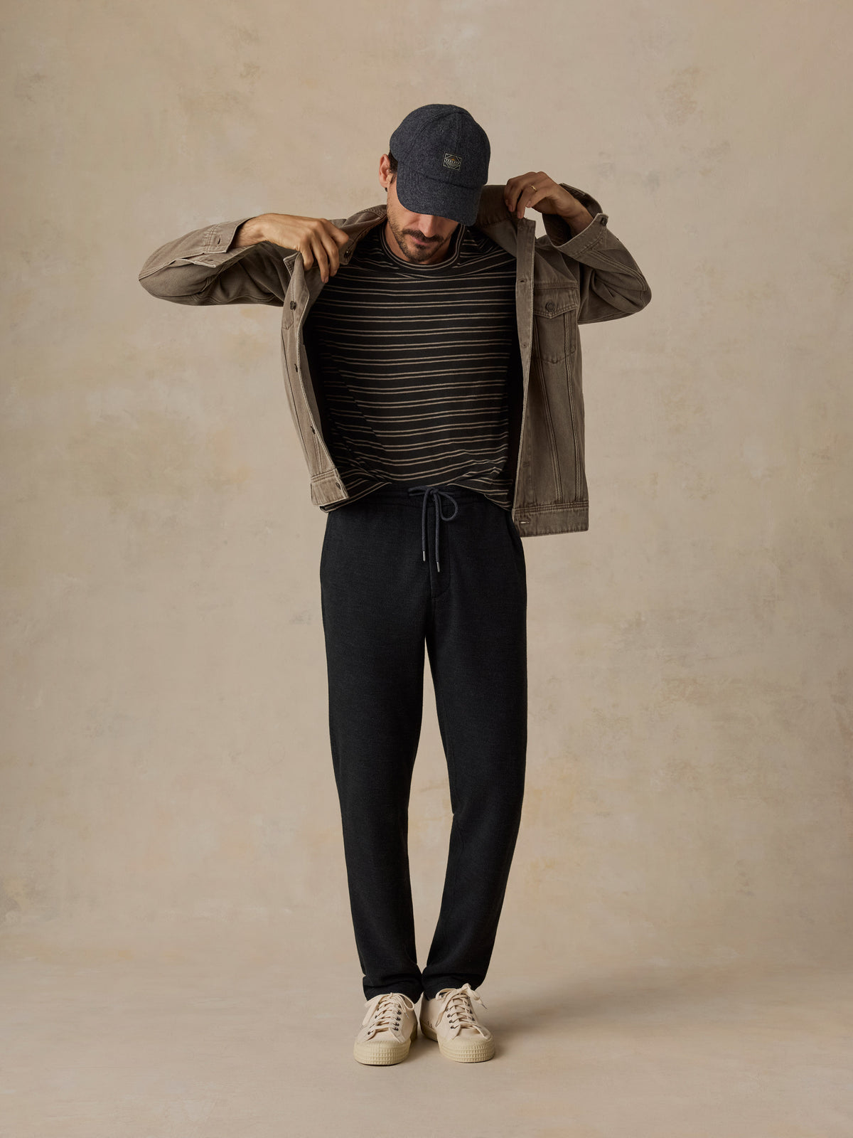 A man modeling a casual outfit consisting of a dark olive green utility jacket, a black and beige striped t-shirt, black sweatpants, a navy blue baseball cap, and off-white sneakers, set against a textured beige studio backdrop., image 6 of 6