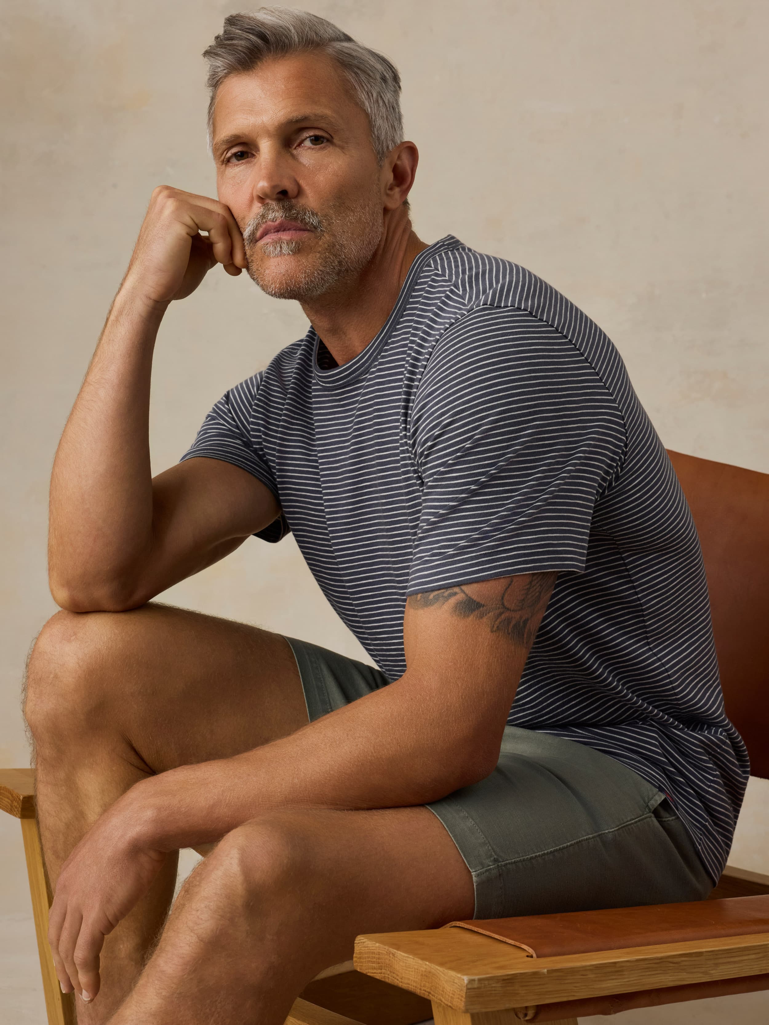 Man sitting on a wooden chair wearing blue striped shirt and green shorts against a beige background