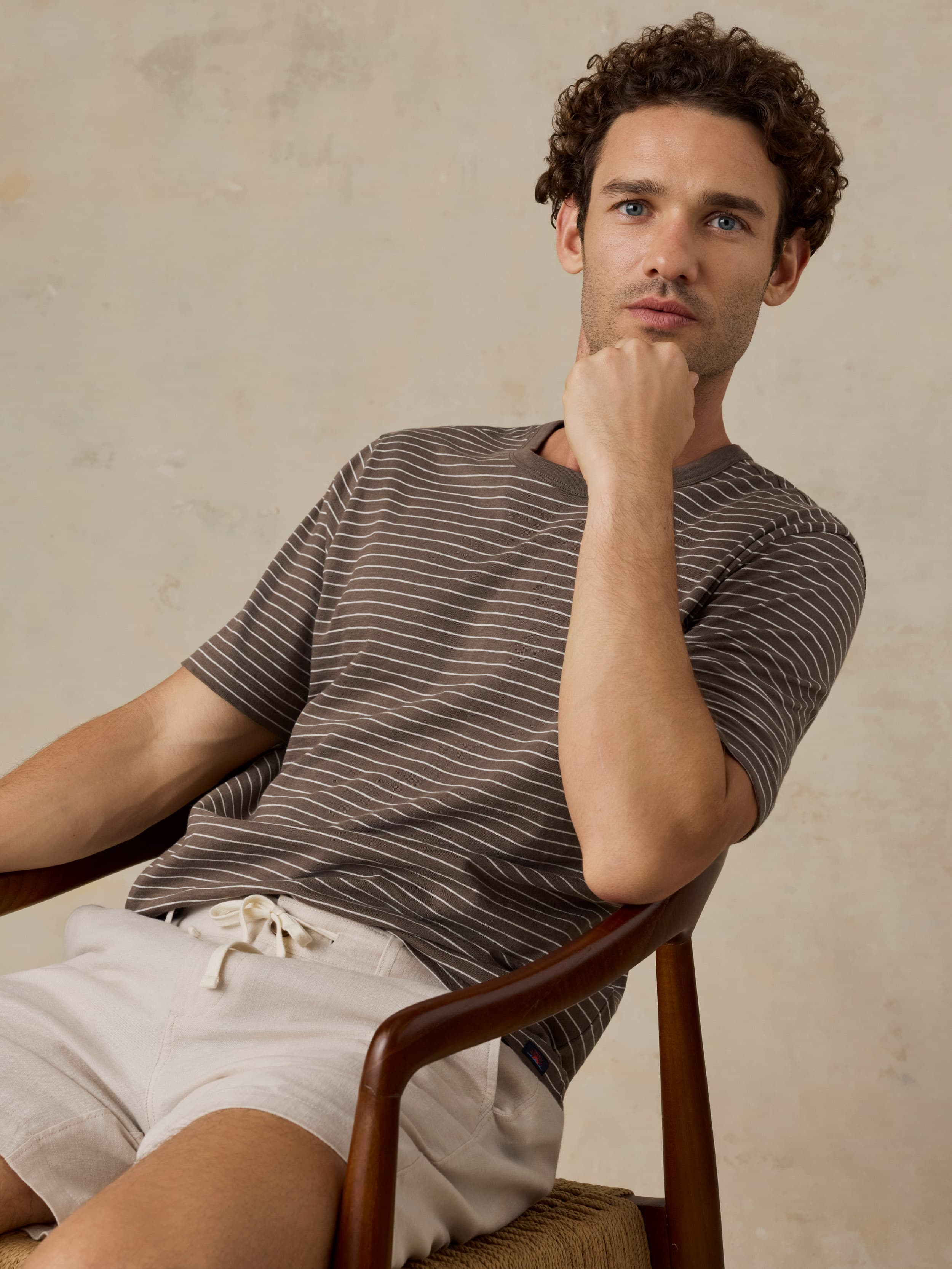 Man sitting in a wooden chair wearing a brown striped t-shirt and beige shorts against a beige background