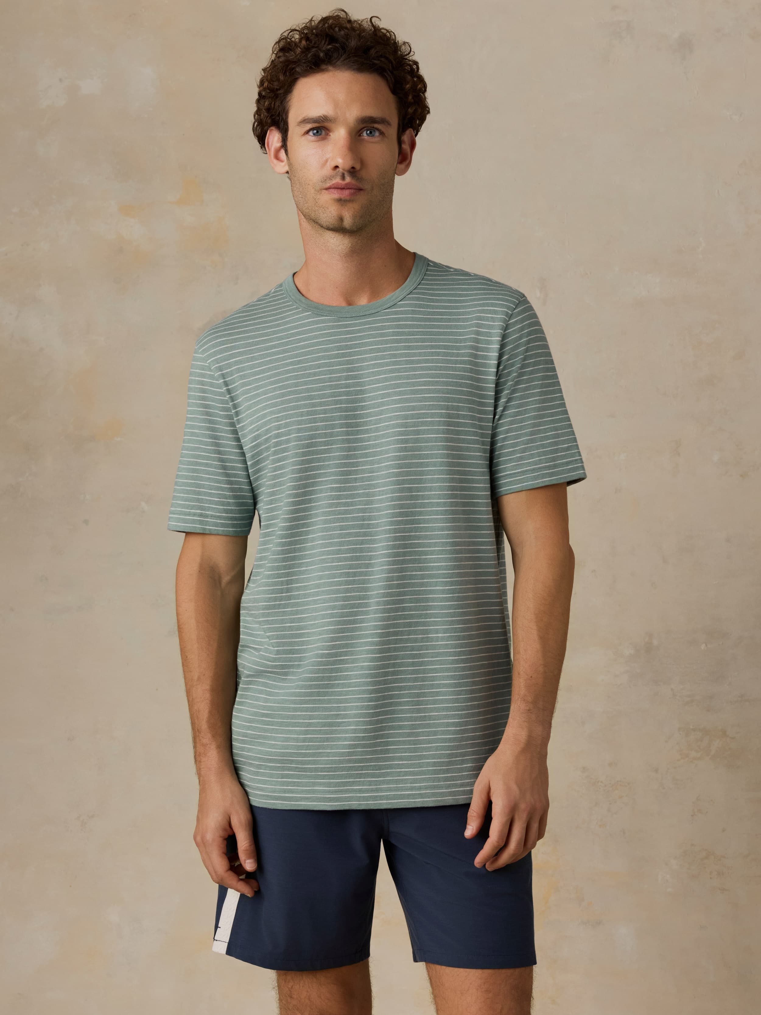 Man wearing a green and white striped tee with navy swim shorts against a beige background