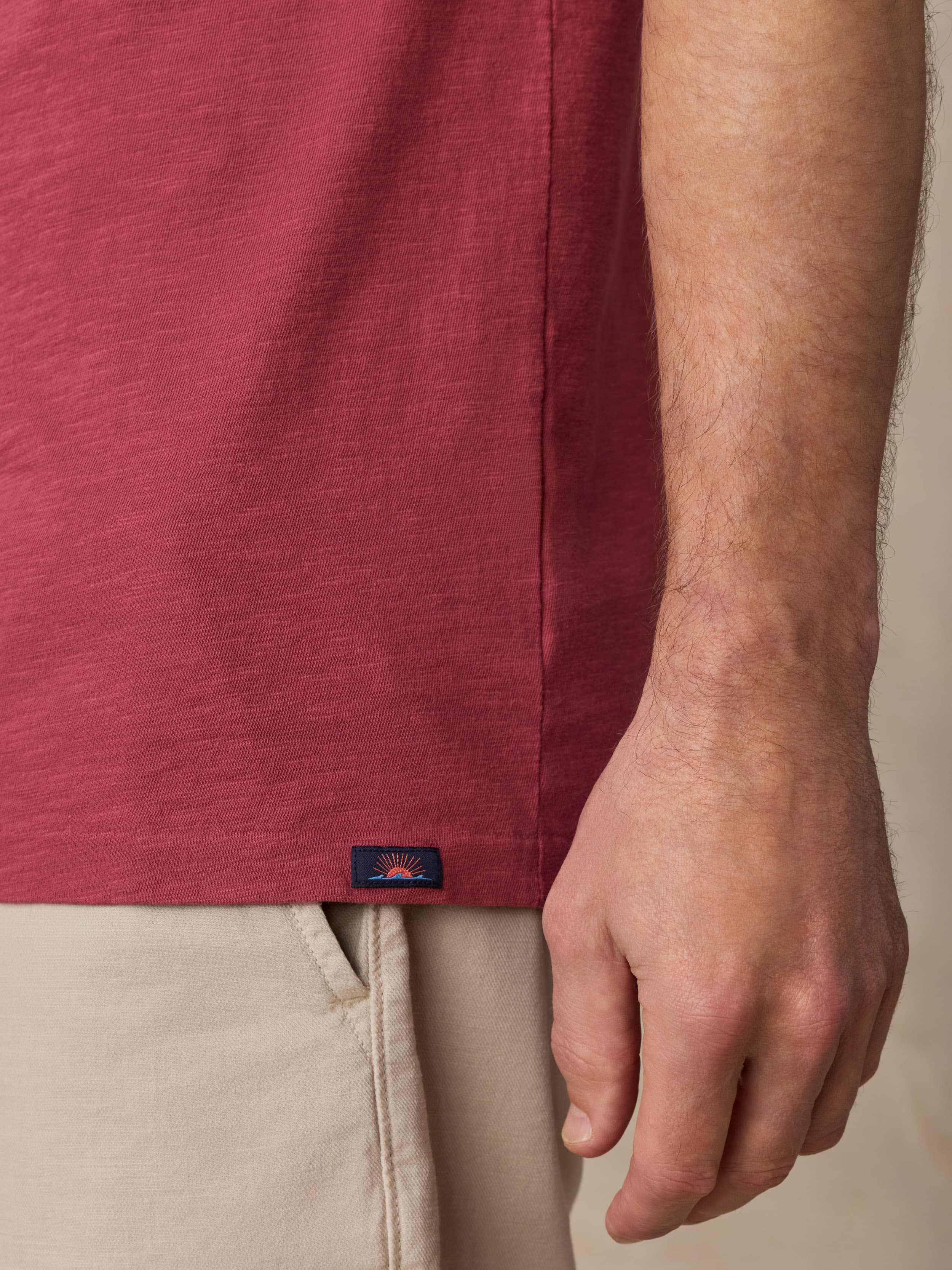 Close-up of a person wearing a red shirt with a Faherty brand logo at the hem and beige shorts.