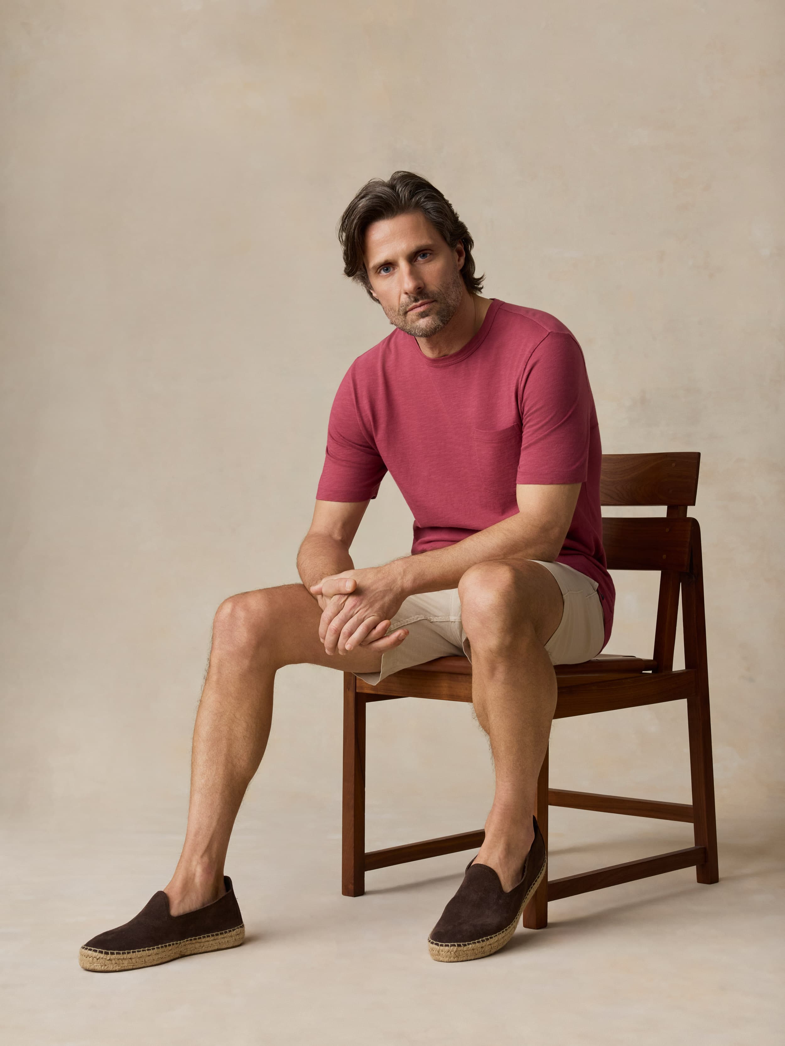 Man sitting on a wooden chair wearing a red t-shirt and beige shorts against a beige background