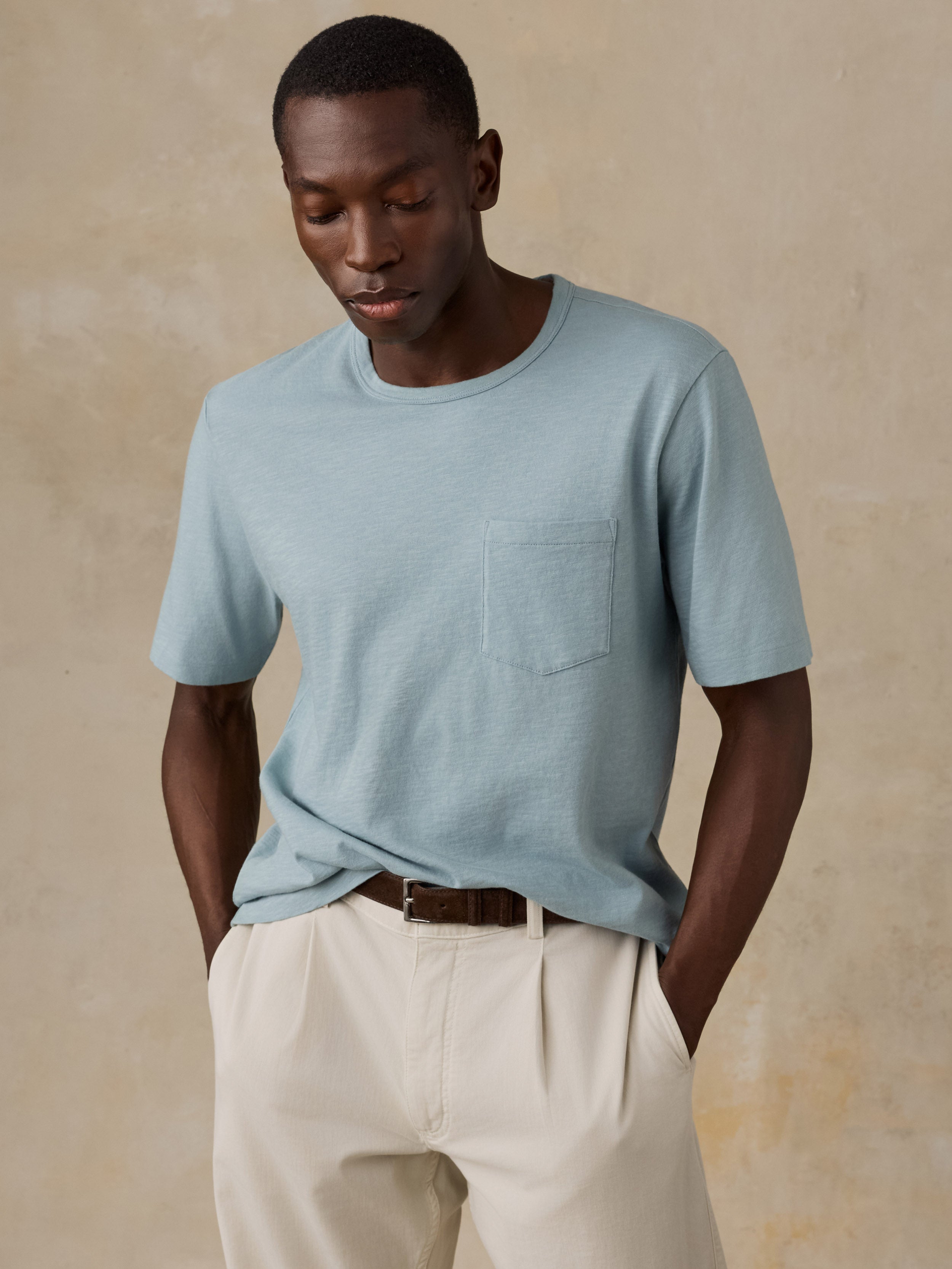 A man wearing a light blue crew-neck short-sleeve knit t-shirt with a chest pocket, tucked into cream-colored pleated trousers, standing against a textured beige background.