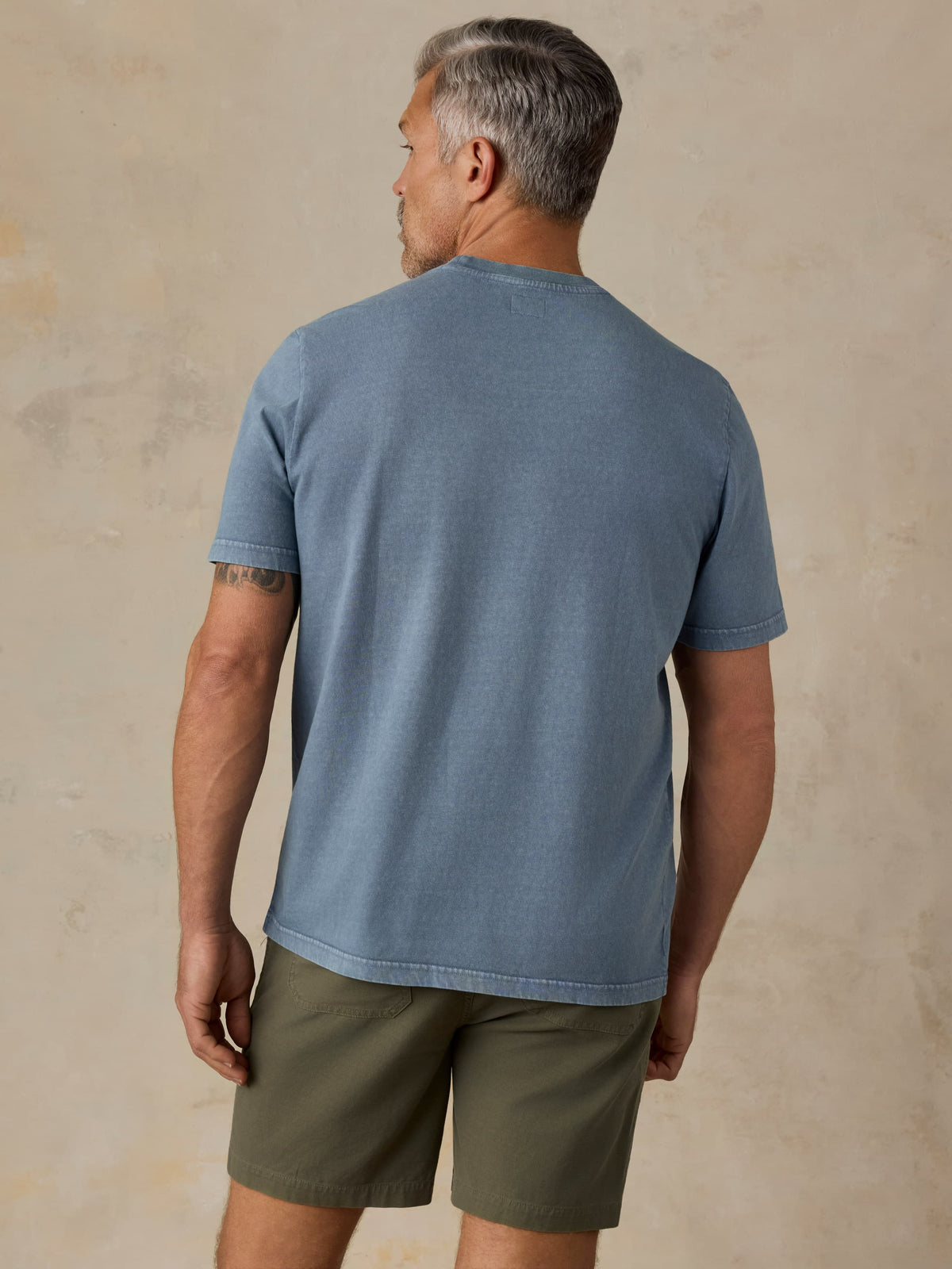 Man wearing a blue pocket tee and green shorts against a beige background, image 4 of 5