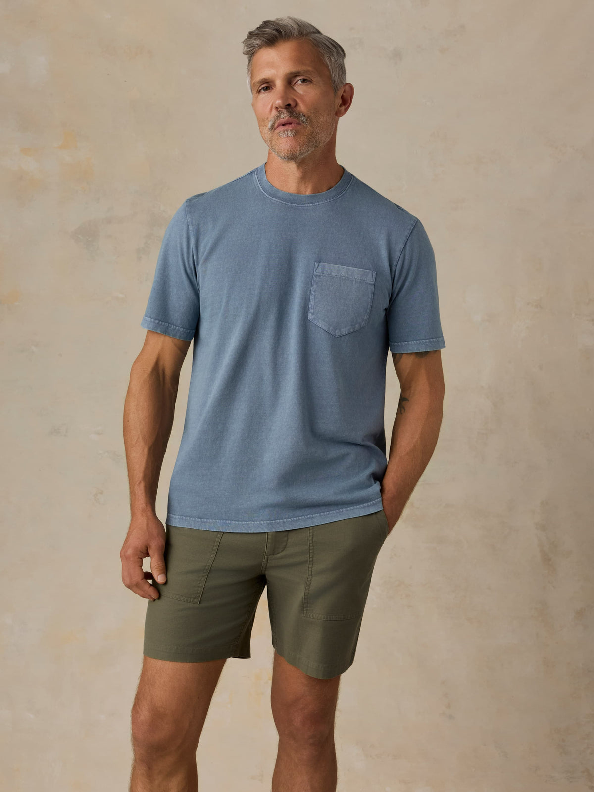 Man wearing a blue pocket tee and green shorts against a beige background, image 1 of 5