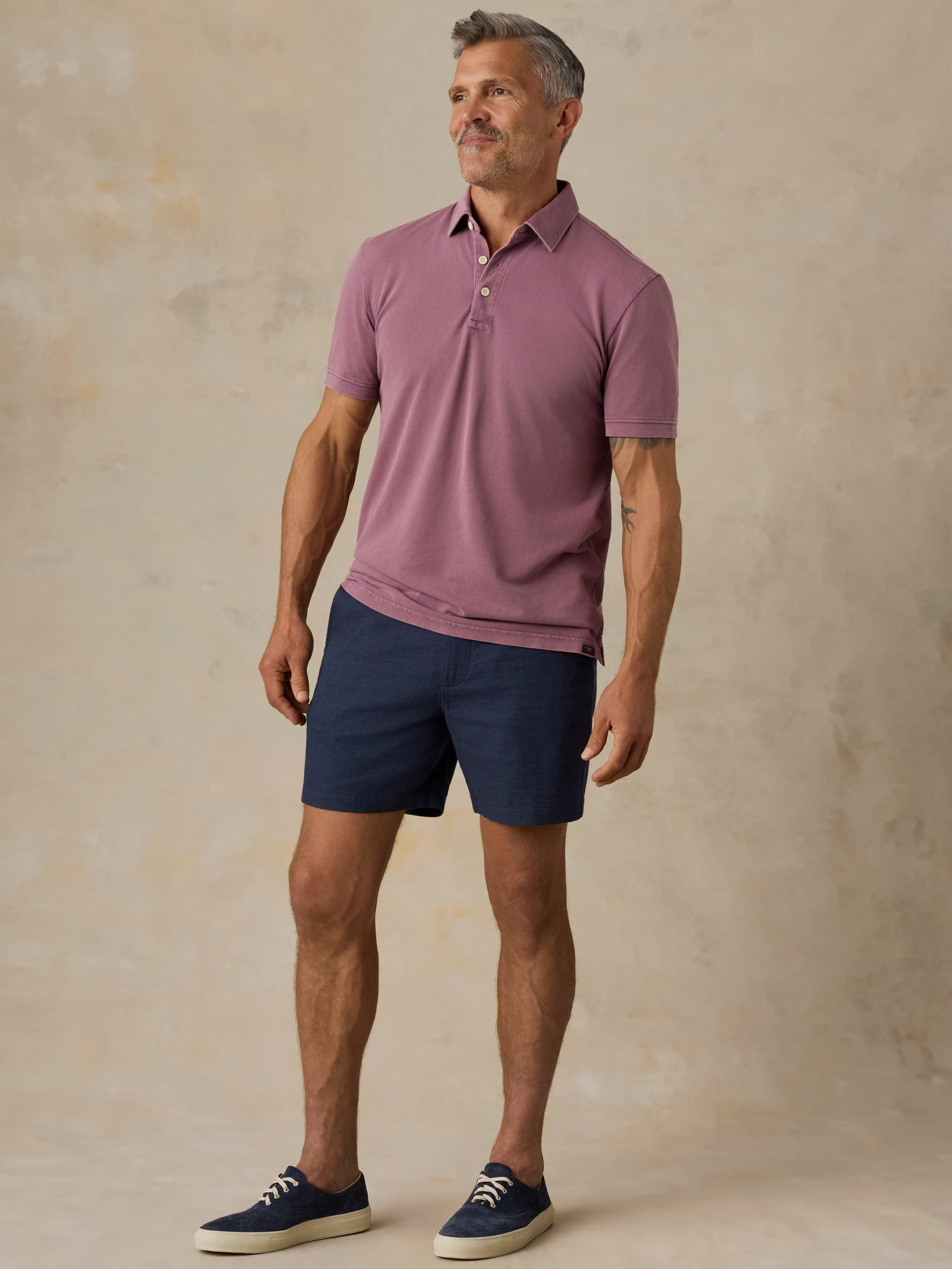 Man wearing a purple polo shirt and navy shorts against a beige background