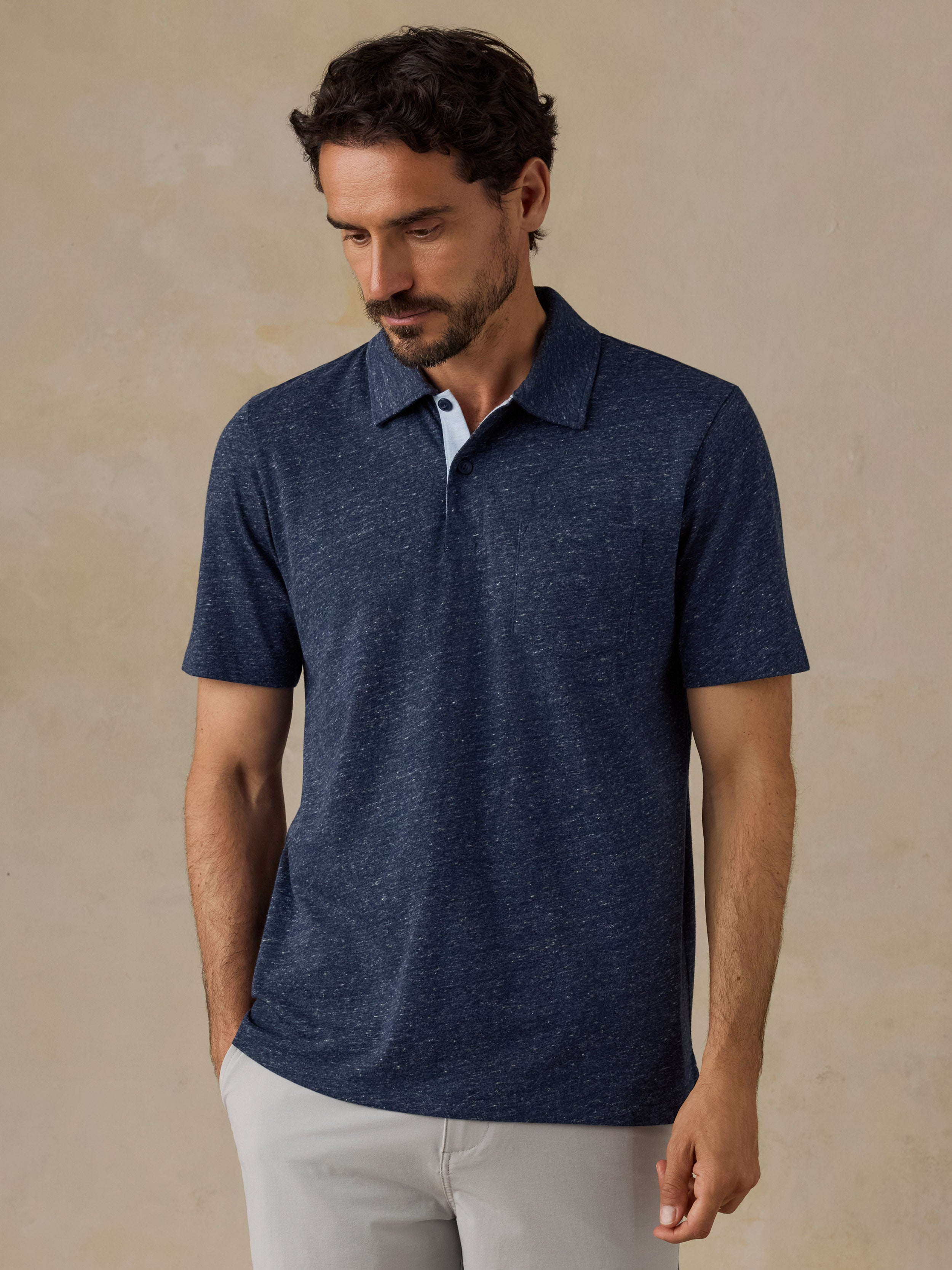 A man wearing a short-sleeve polo shirt in a heathered navy blue color, paired with light gray trousers, standing against a textured beige wall.