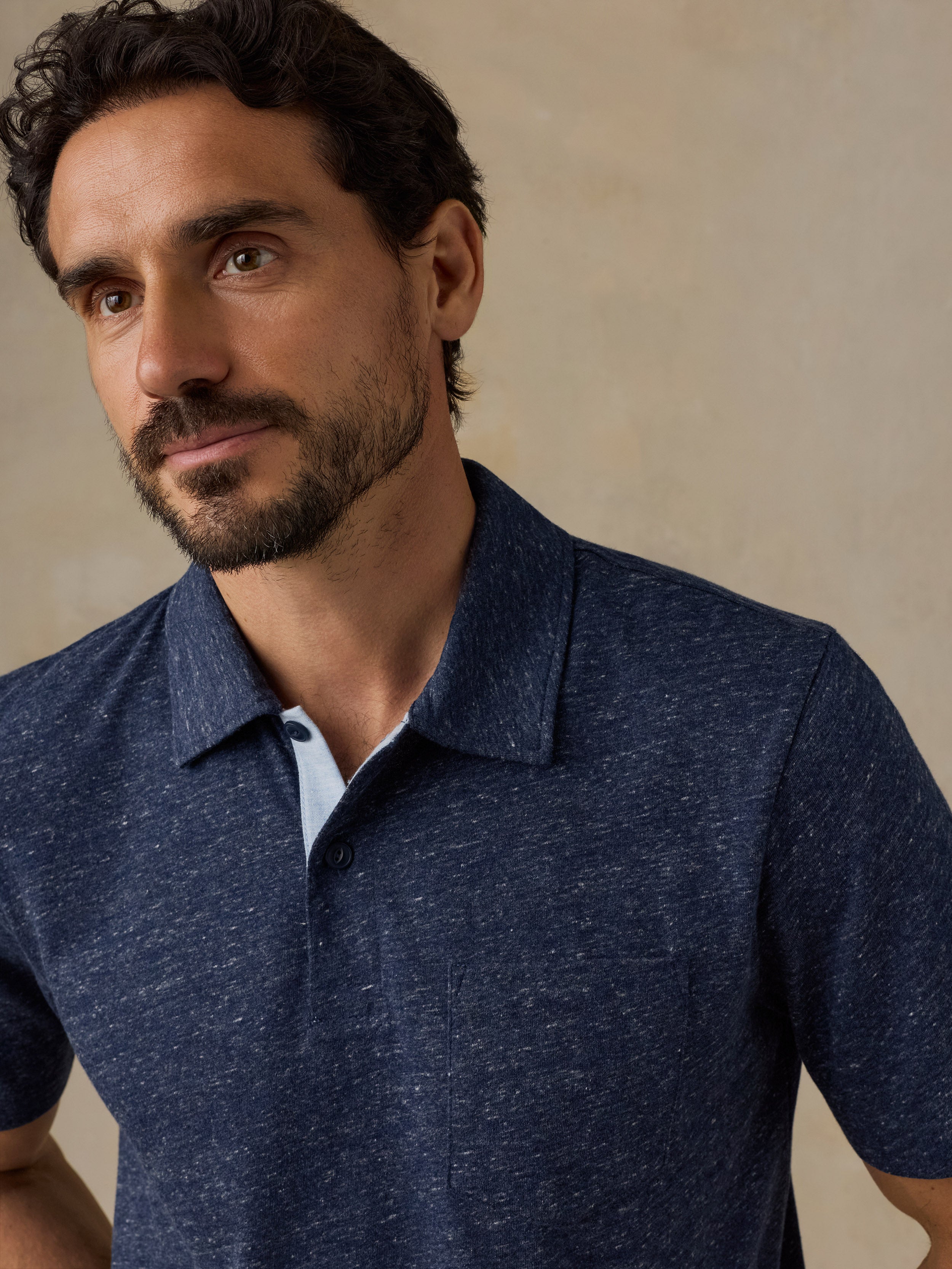 A close-up of a man wearing a dark heathered blue short-sleeve polo shirt against a neutral beige background.