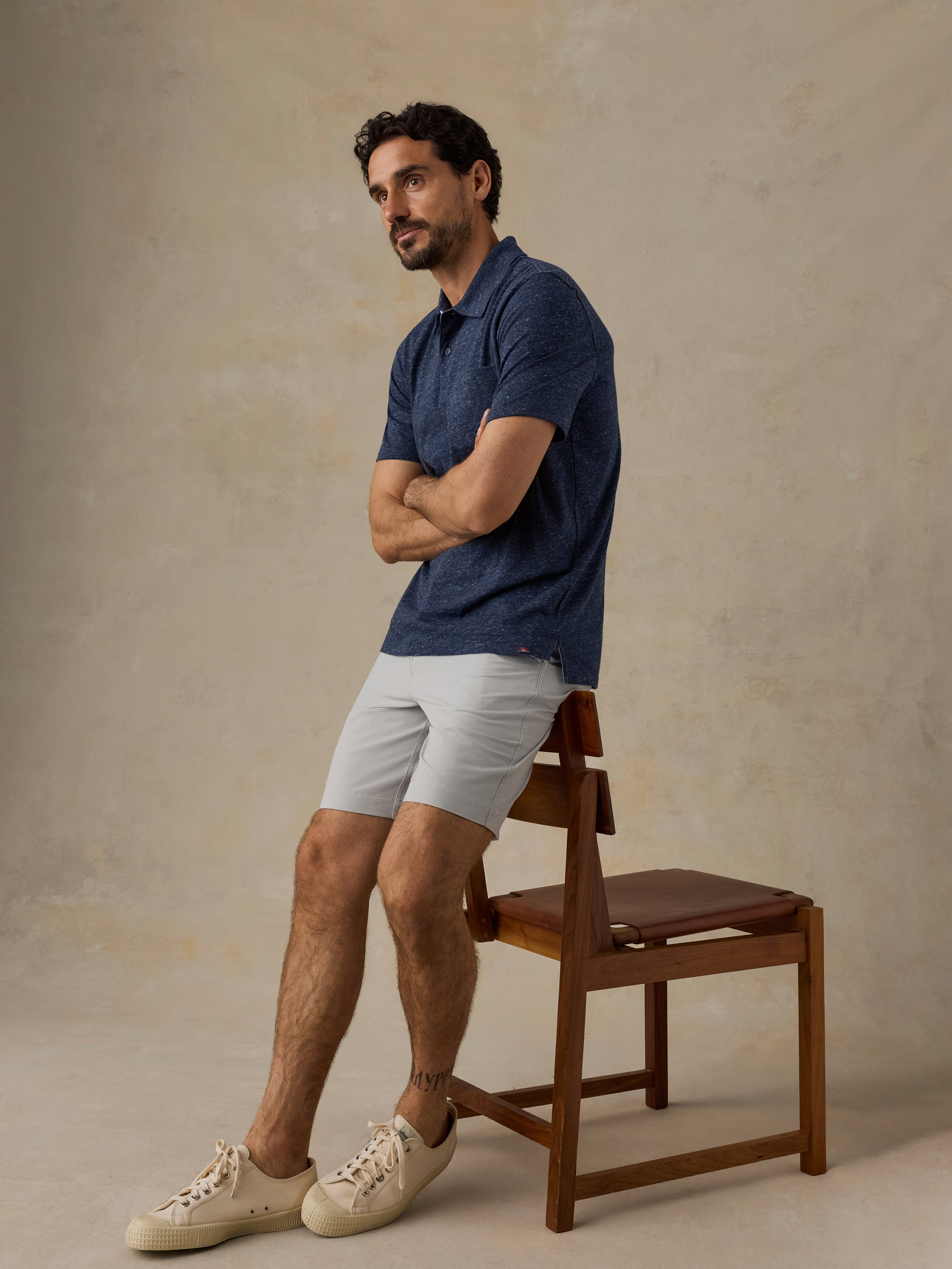 Man wearing a blue polo shirt and beige shorts sitting on a chair against a beige background