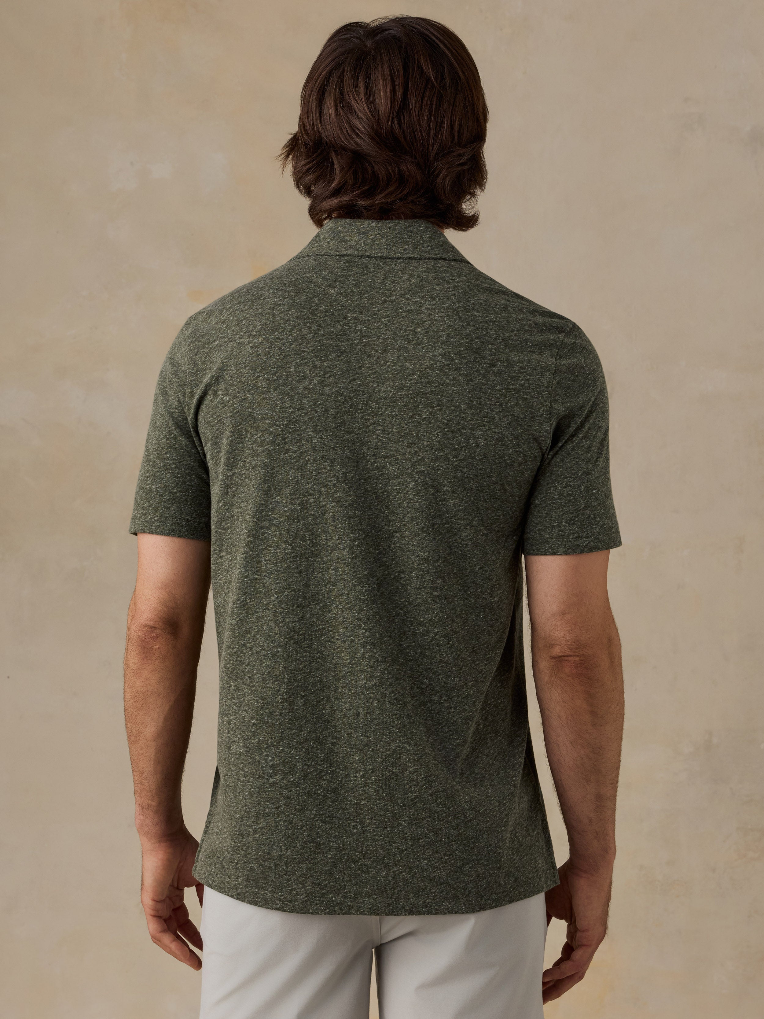 A man viewed from the back wearing a short-sleeve knit polo shirt in heathered olive green, paired with light-colored pants, against a textured beige studio backdrop.