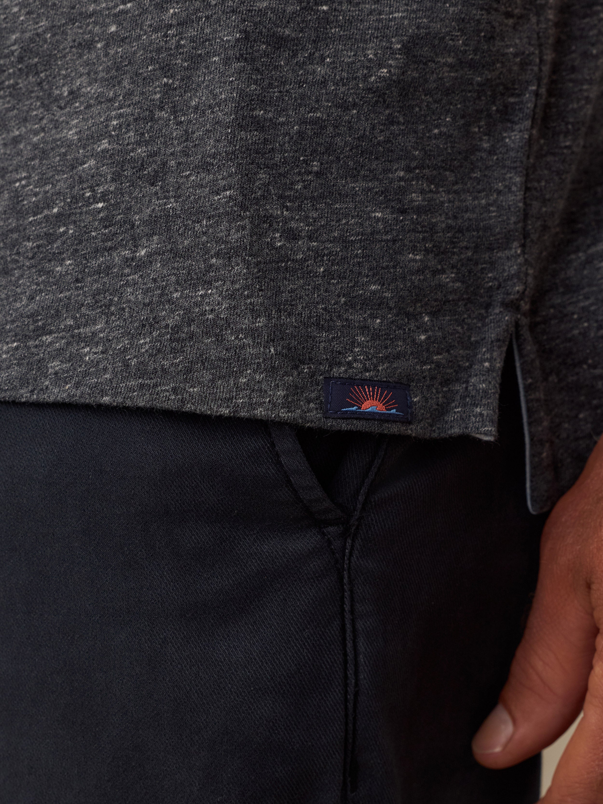 Close-up of a dark heather gray knit top worn with black pants, featuring a small Faherty brand patch near the hem and part of a man hand visible on the right.