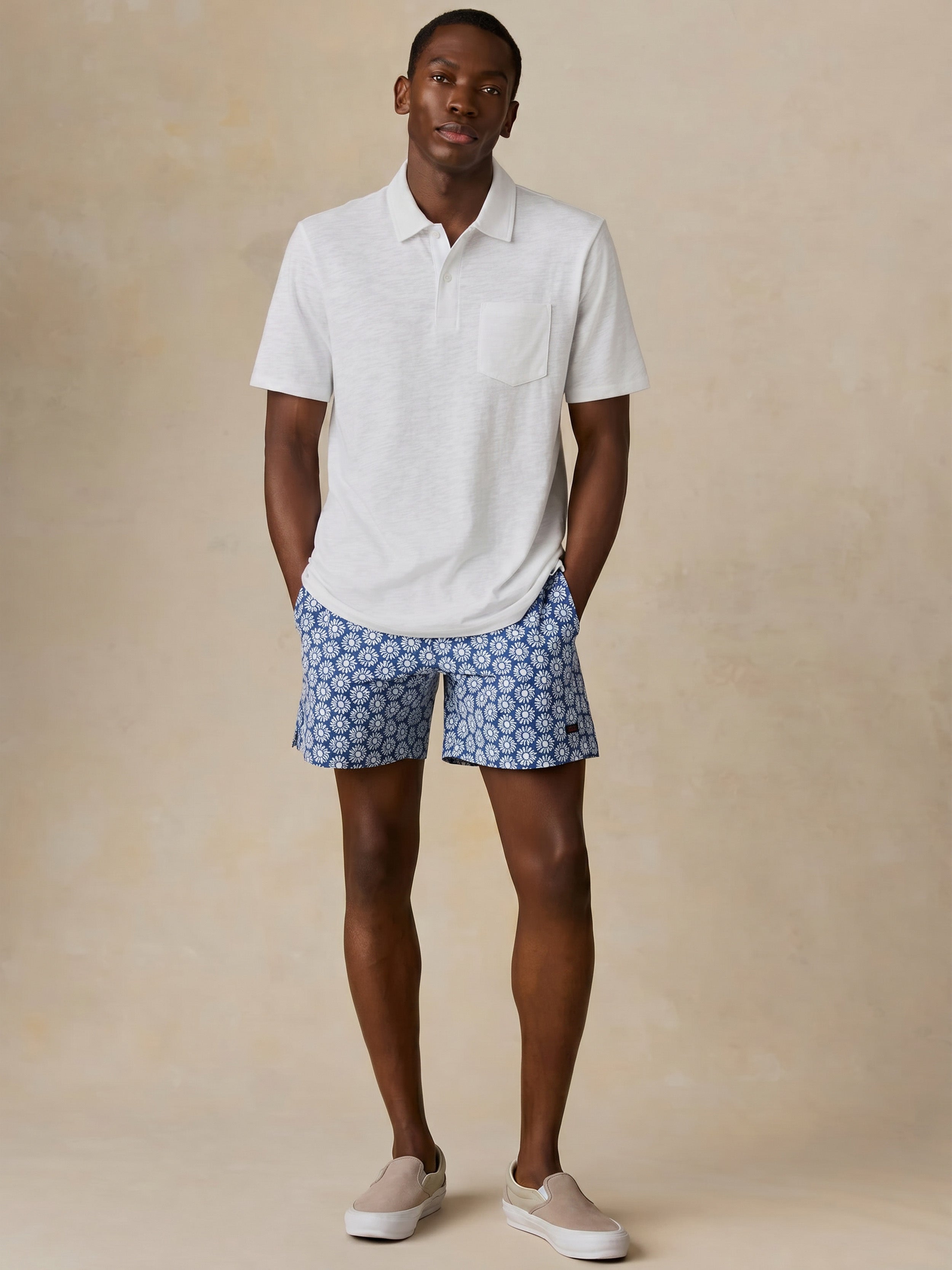 Man wearing a white short sleeve polo and blue printed swim shorts against a beige background