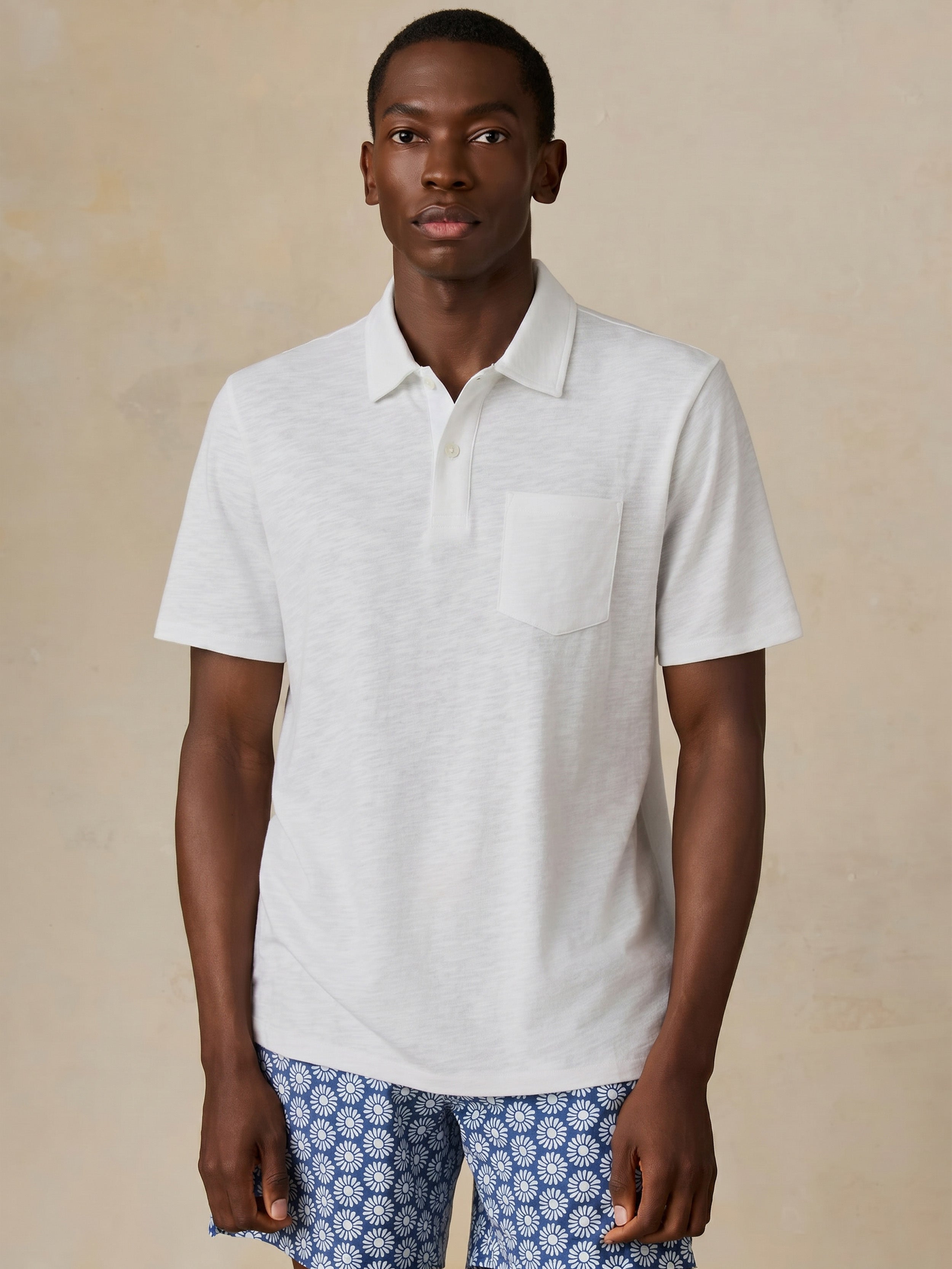Man wearing a white short sleeve polo and blue printed swim shorts against a beige background