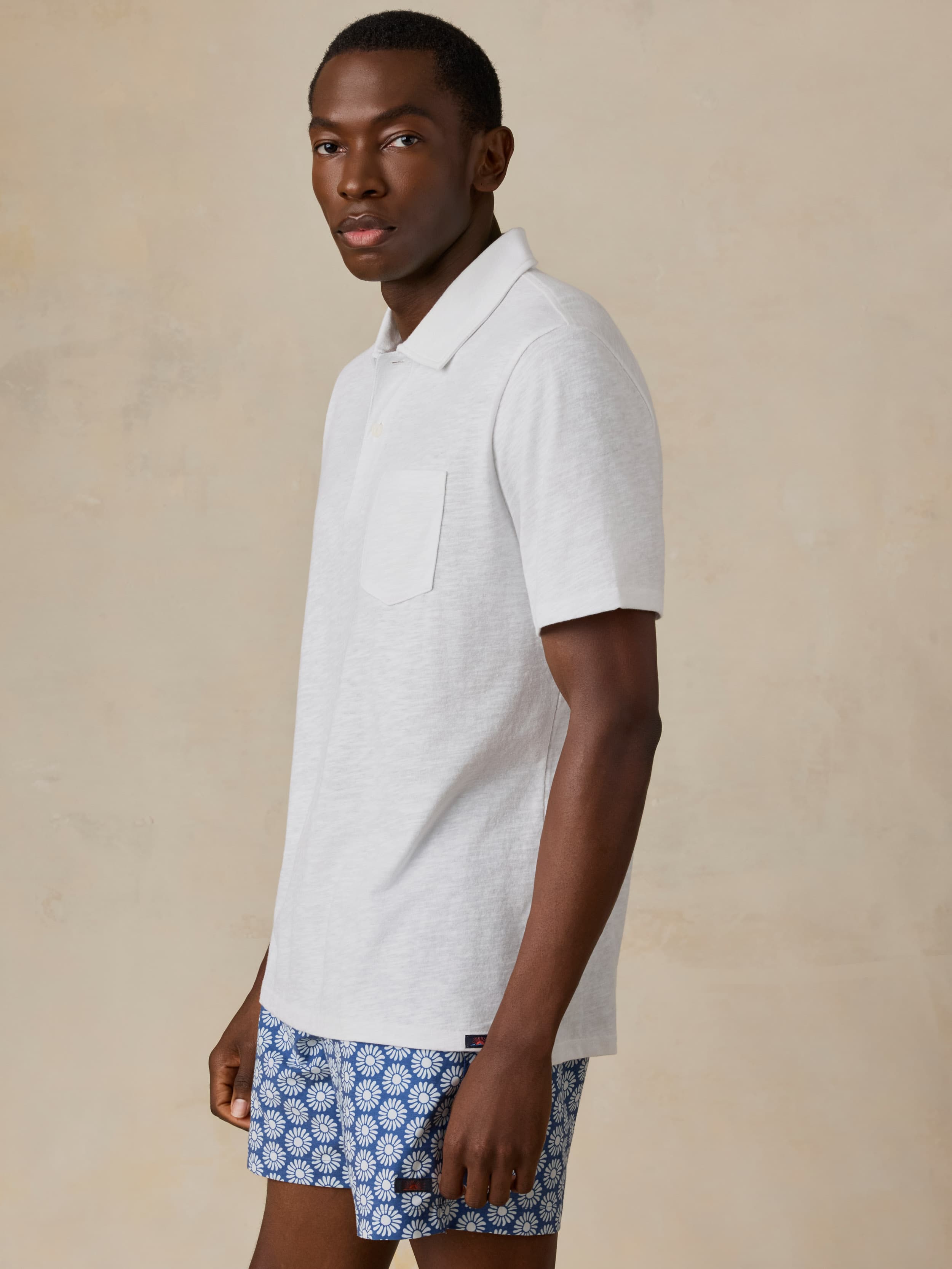 Man wearing a white short sleeve polo and blue printed swim shorts against a beige background