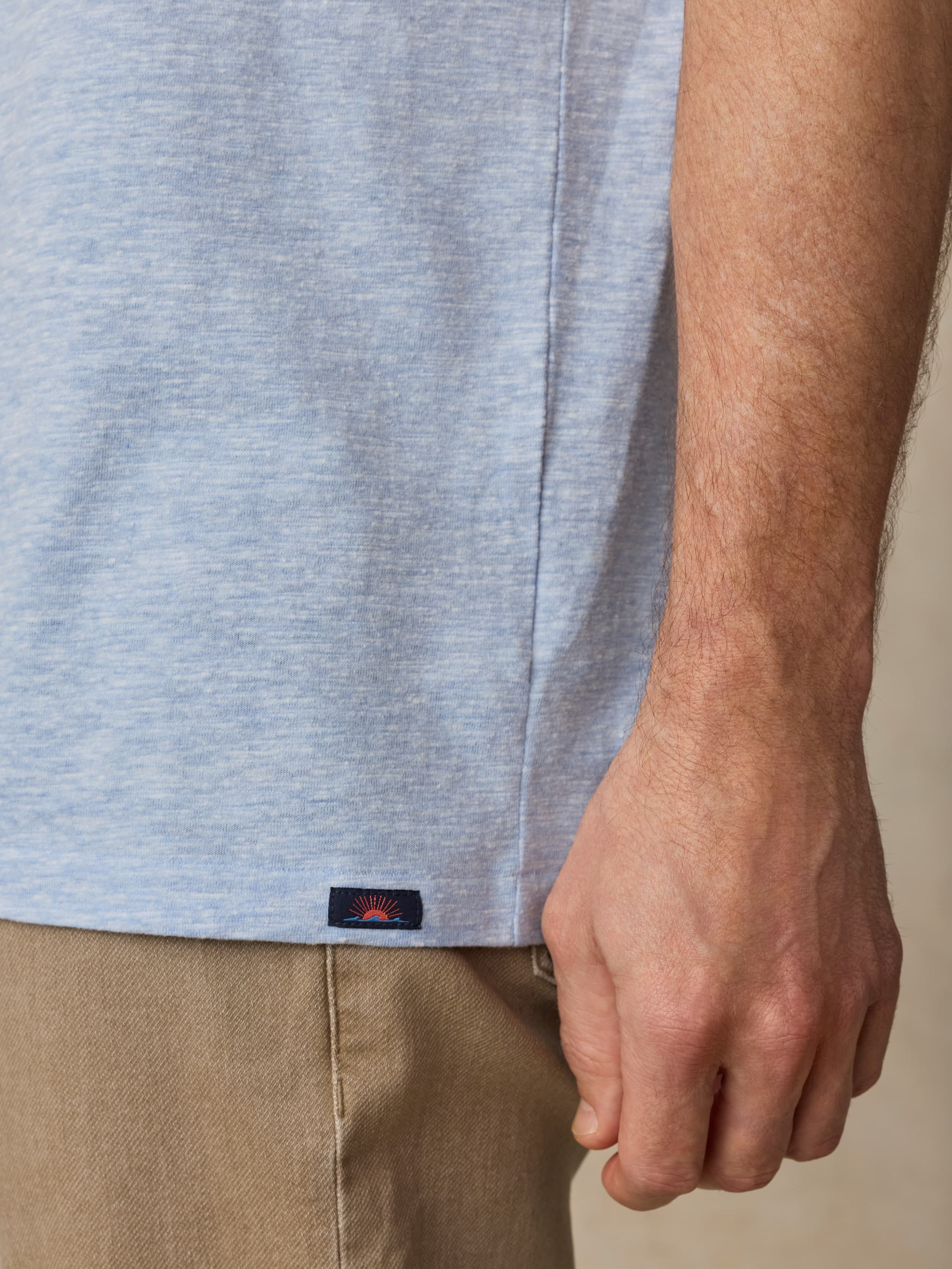 Close-up of a man wearing a light blue t-shirt with visible Faherty brand label near the hem and beige pants against a beige background