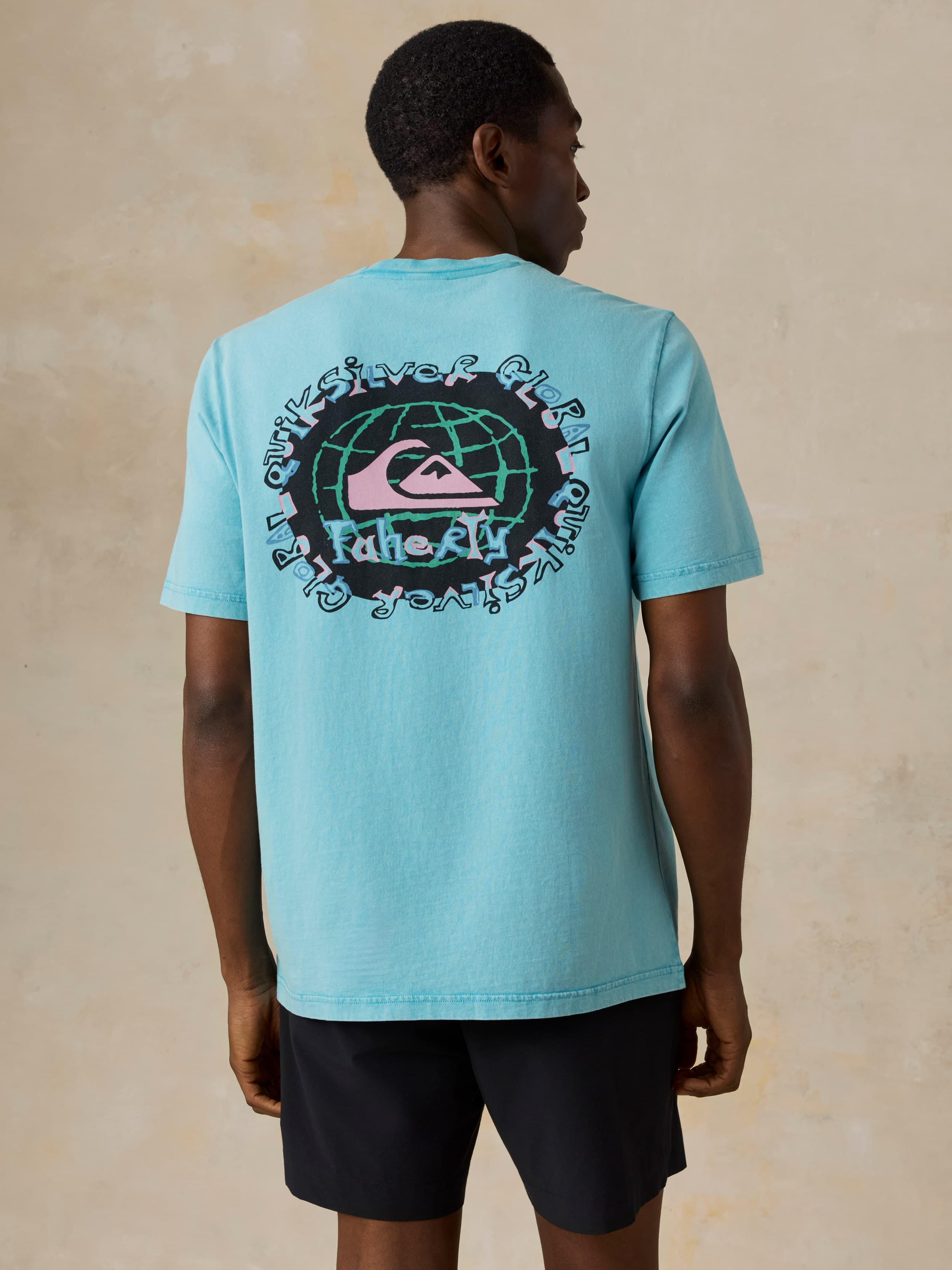 Man wearing a blue Faherty x Quiksilver tee and black shorts against a beige background