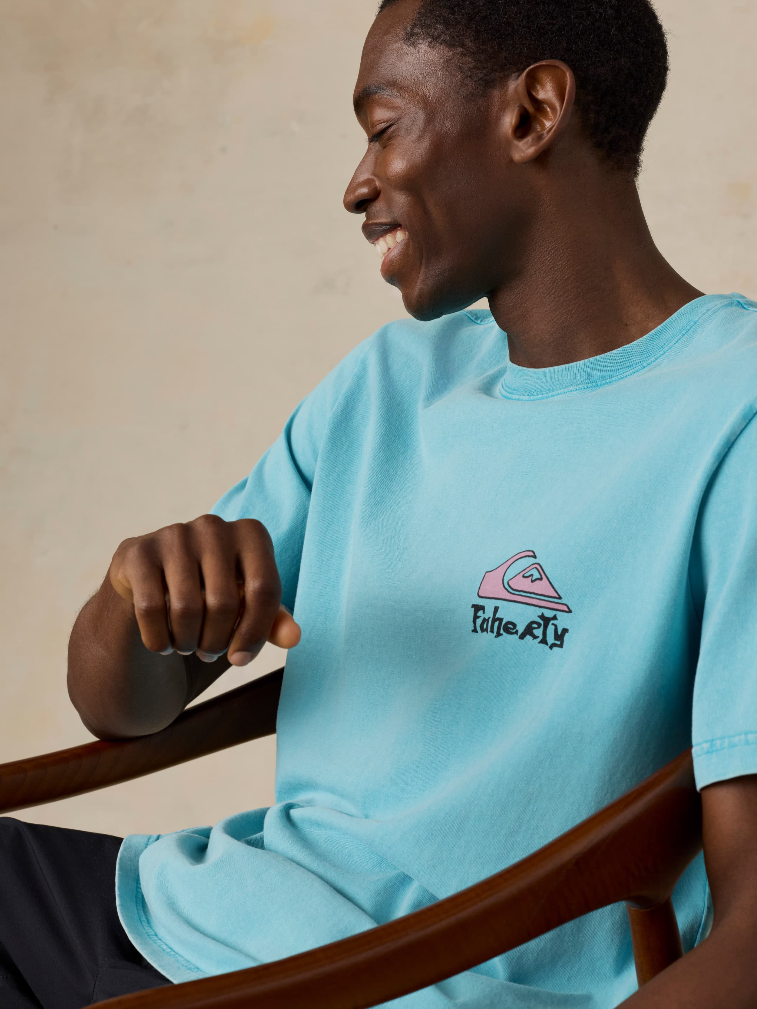 Man sitting in a chair wearing a blue Faherty x Quiksilver tee and black shorts against a beige background