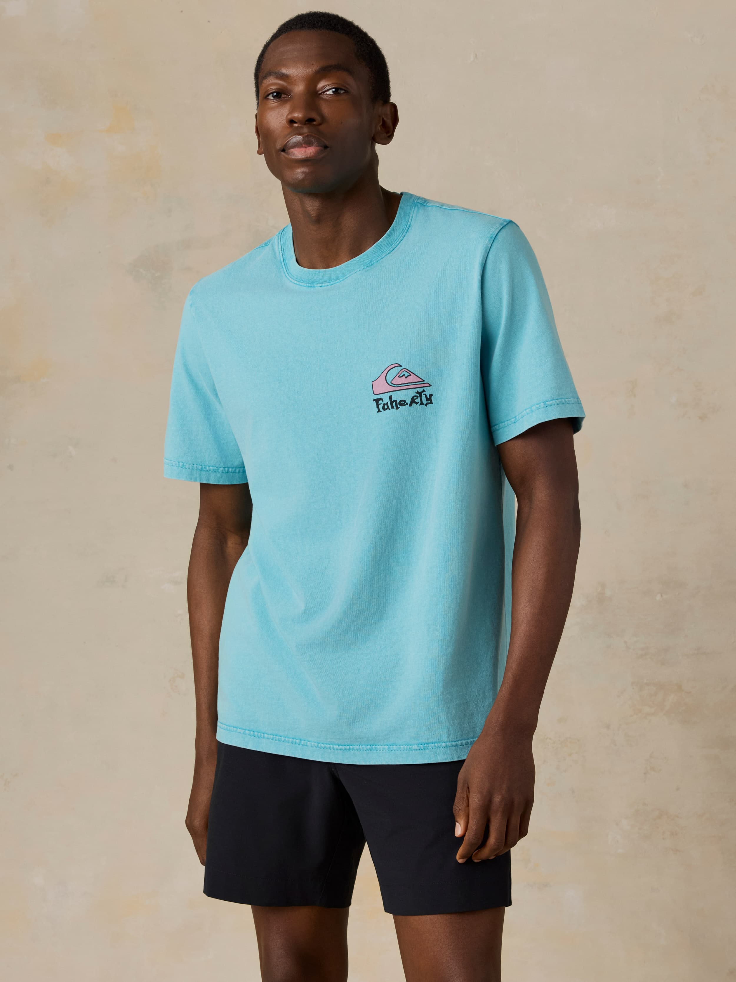 Man wearing a blue Faherty x Quiksilver tee and black shorts against a beige background