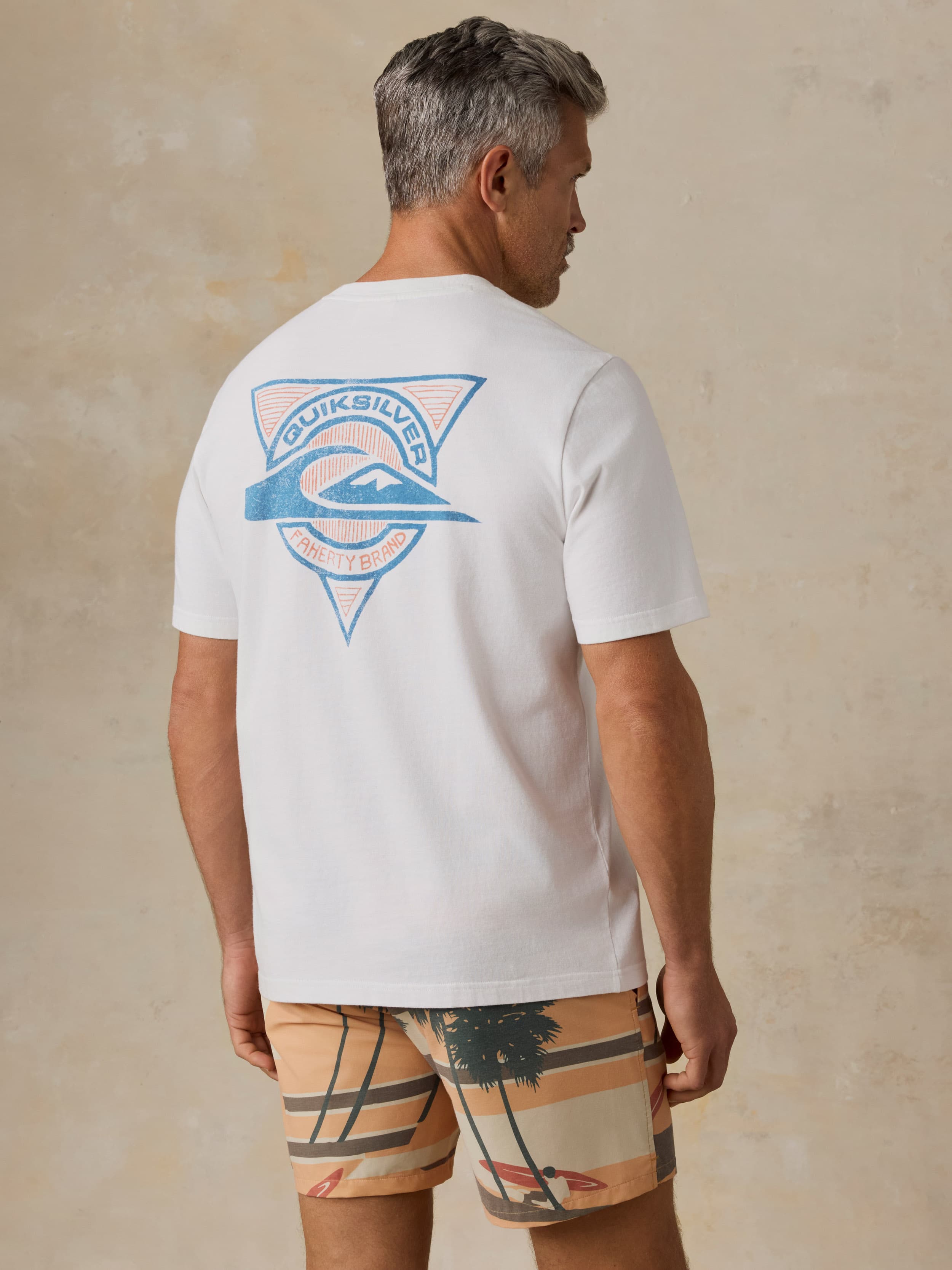 Man wearing a white Faherty x Quiksilver t-shirt with printed swim trunks against a beige background