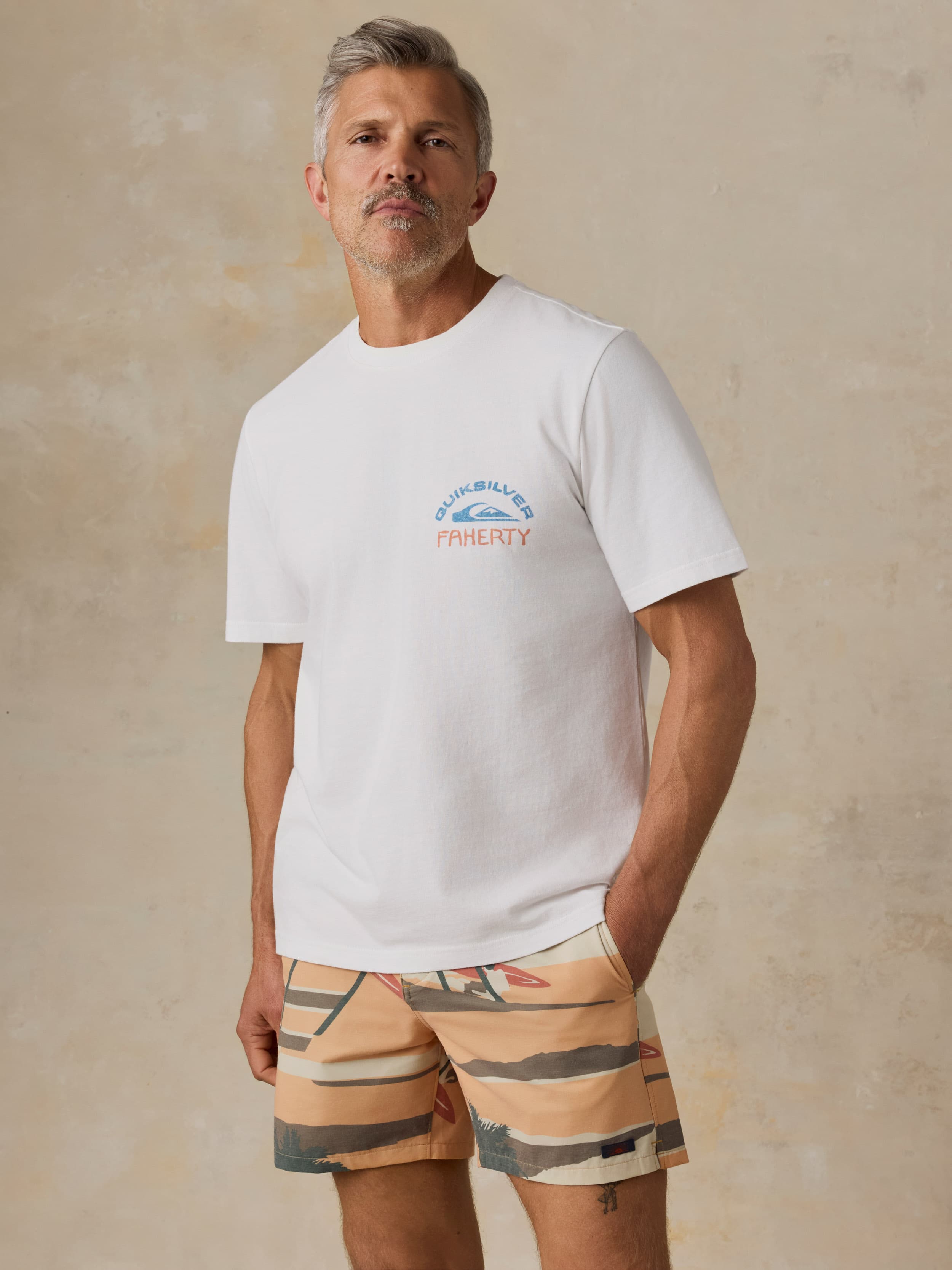Man wearing a white Faherty x Quiksilver t-shirt with printed swim trunks against a beige background