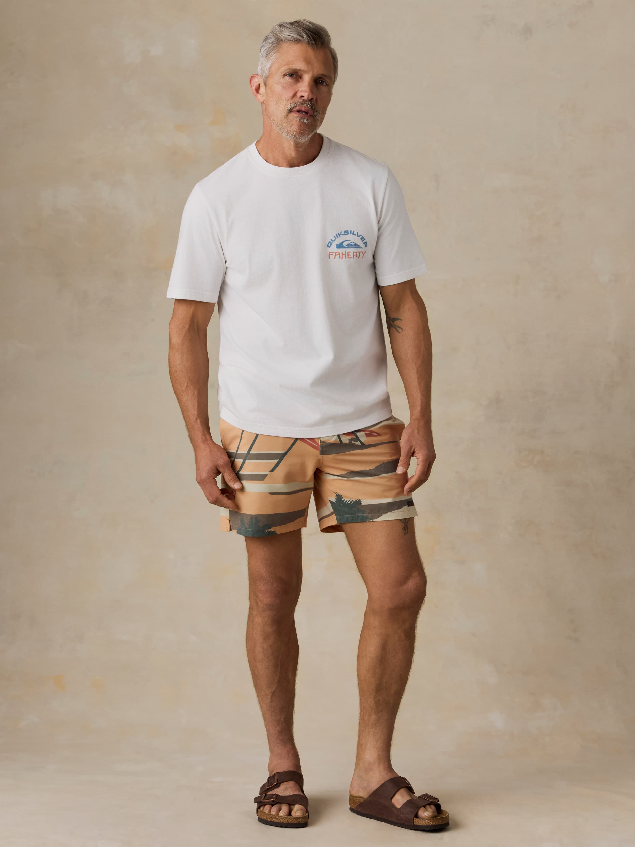 Man wearing a white Faherty x Quiksilver t-shirt with printed swim trunks against a beige background