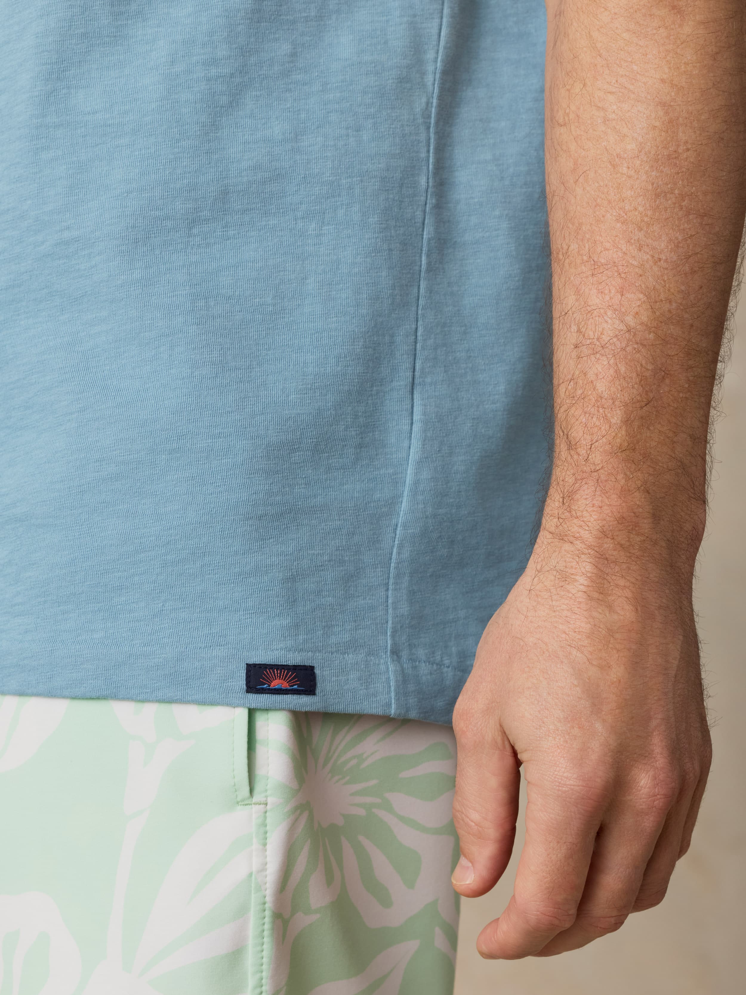 Close-up of a man wearing a light blue shirt with a visible Faherty brand label near the hem and light green shorts with a floral pattern against a beige background