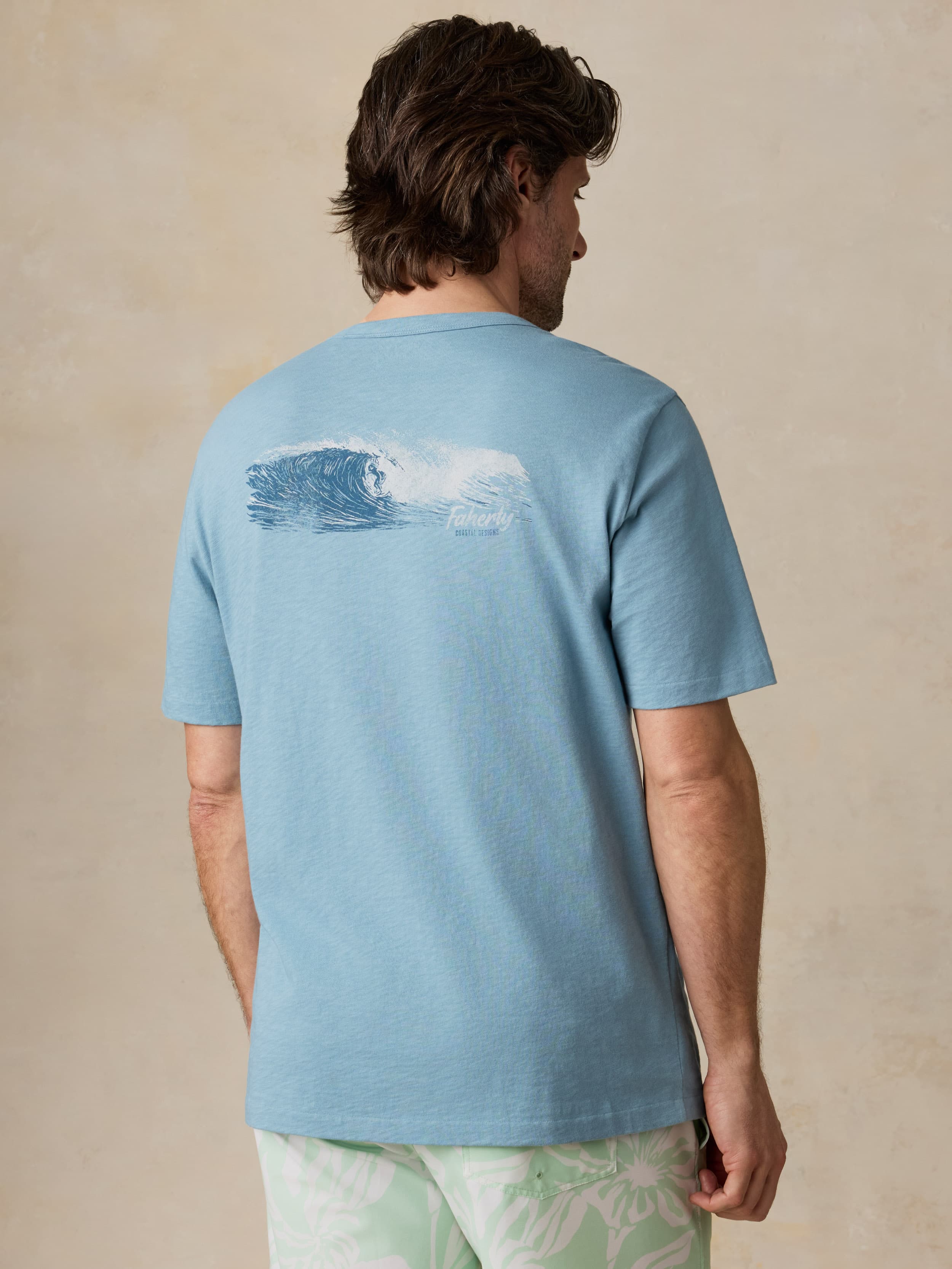 Man wearing a light blue t-shirt with a wave design on the back, standing against a beige background.