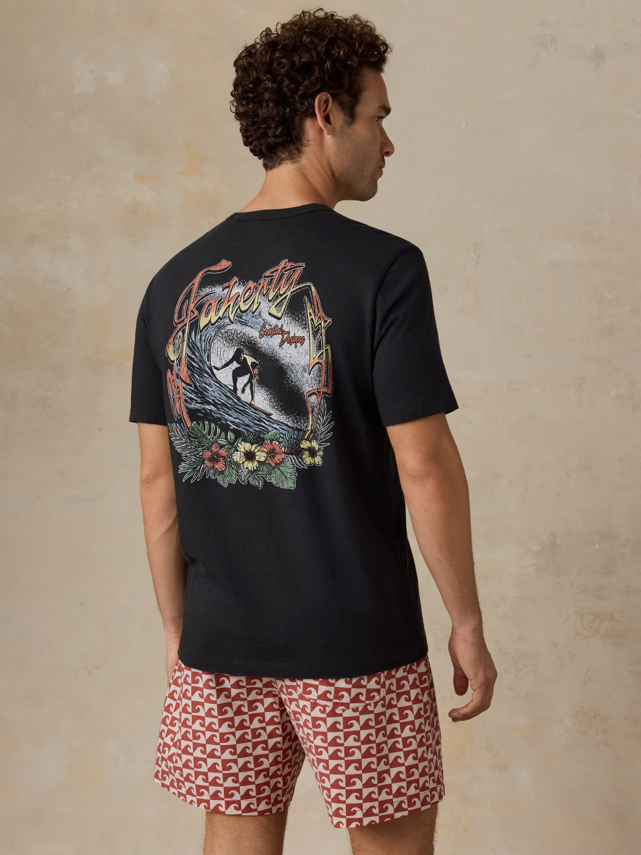 Man wearing a black t-shirt with a colorful graphic on the back and patterned shorts, standing against a beige background.