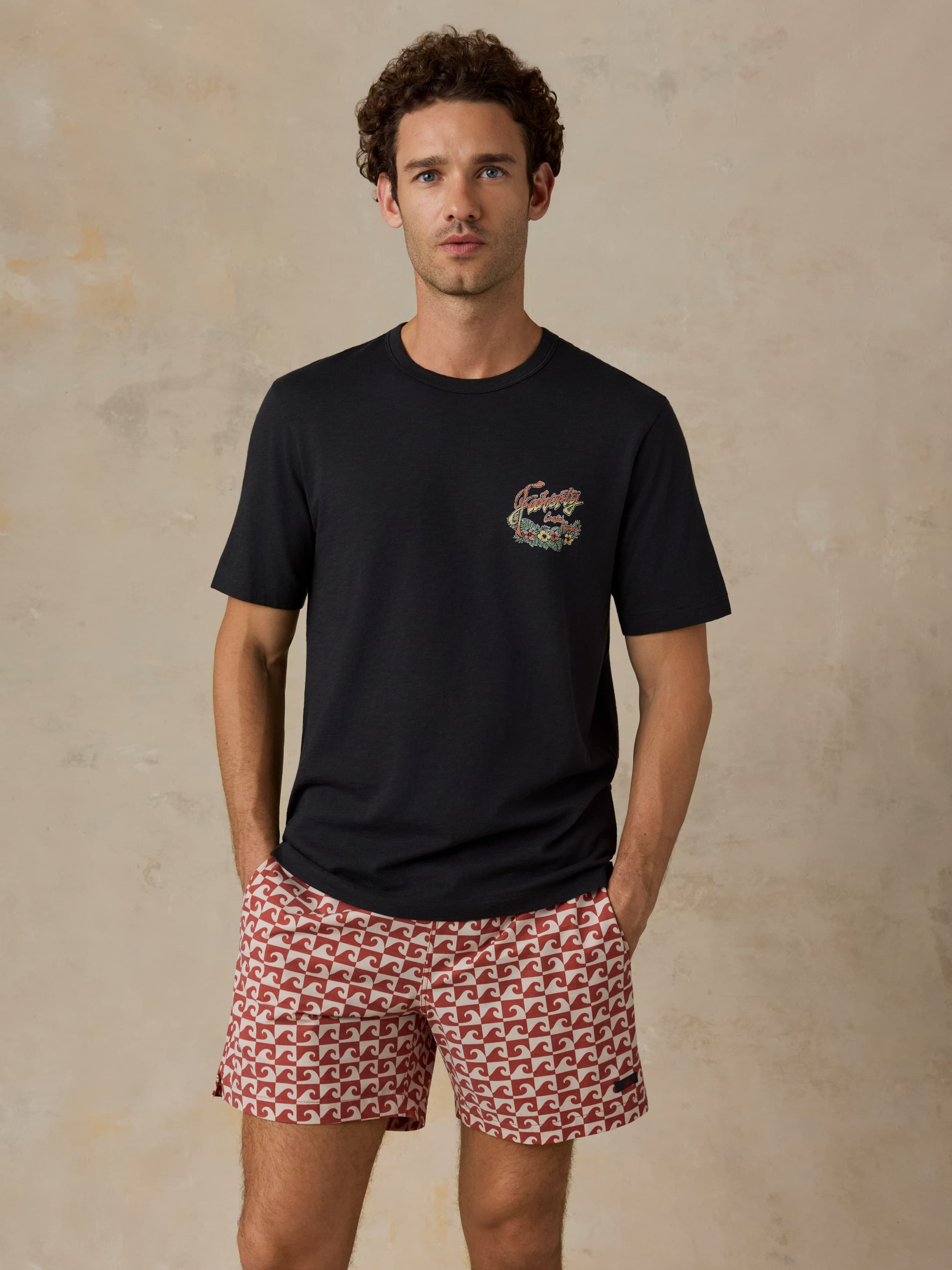 Man wearing a black t-shirt with a logo and red patterned shorts against a beige background