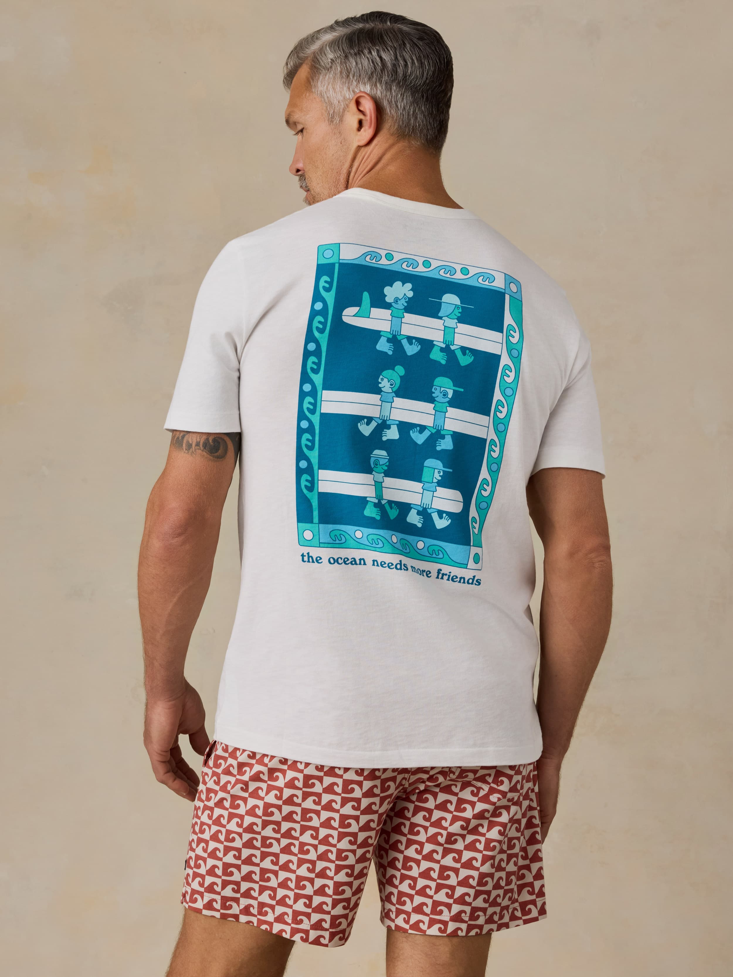 Man wearing a white t-shirt with a Faherty x Surfrider Foundation logo and patterned shorts on a beige background