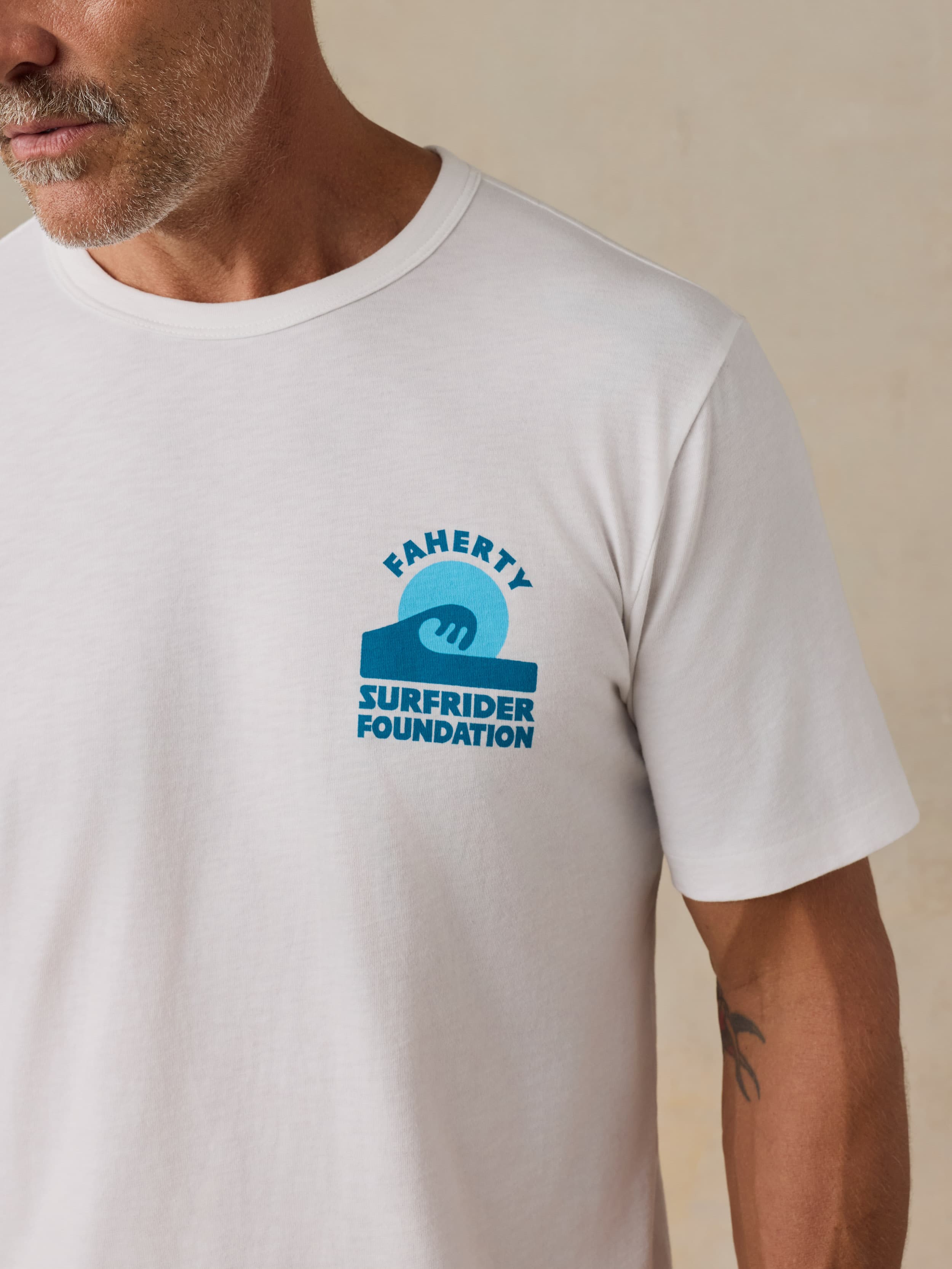 Man wearing a white t-shirt with a Faherty x Surfrider Foundation logo on a beige background