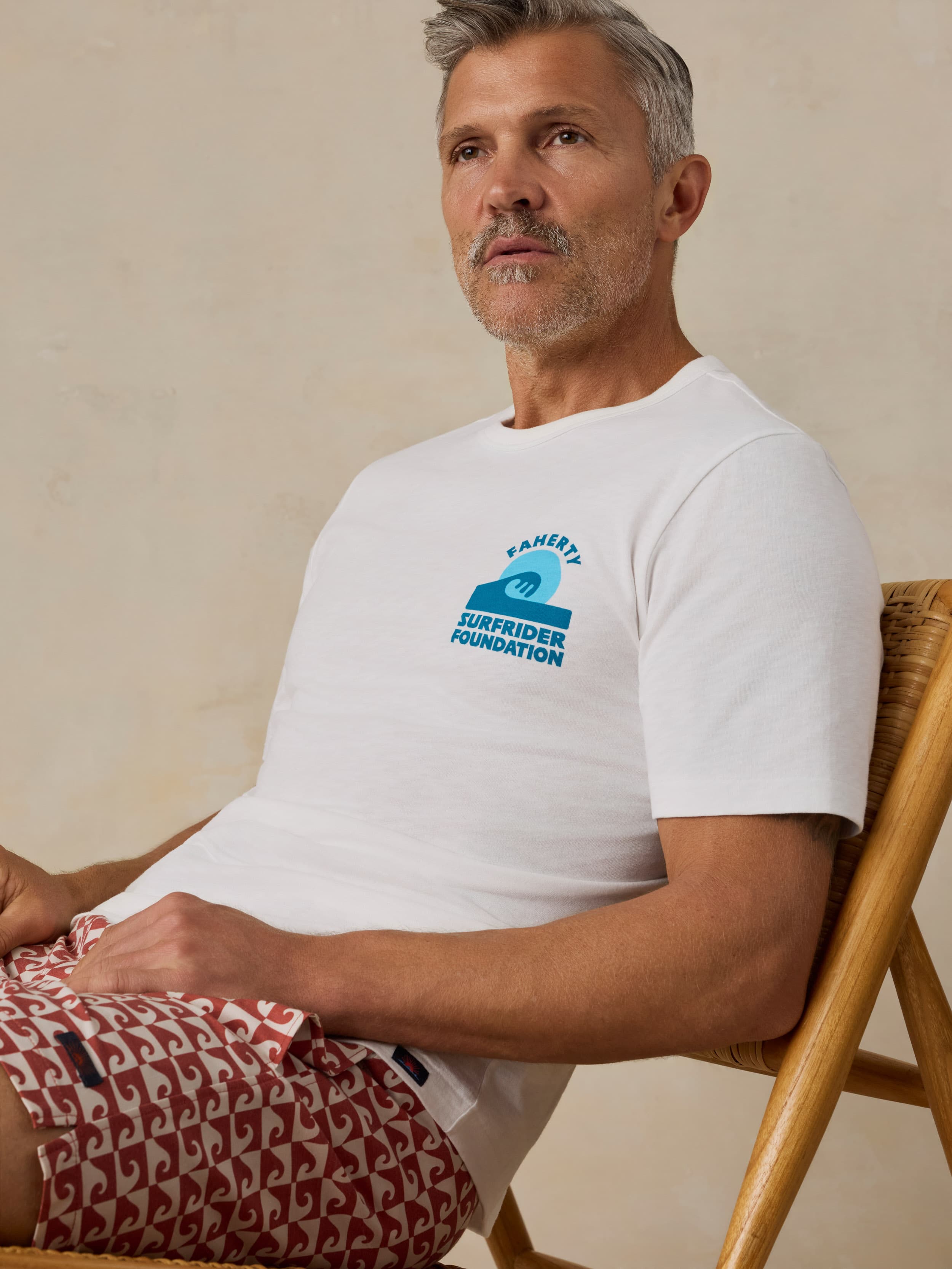Man wearing a white t-shirt with a Faherty x Surfrider Foundation logo and patterned shorts sitting on a wooden chair on a beige background