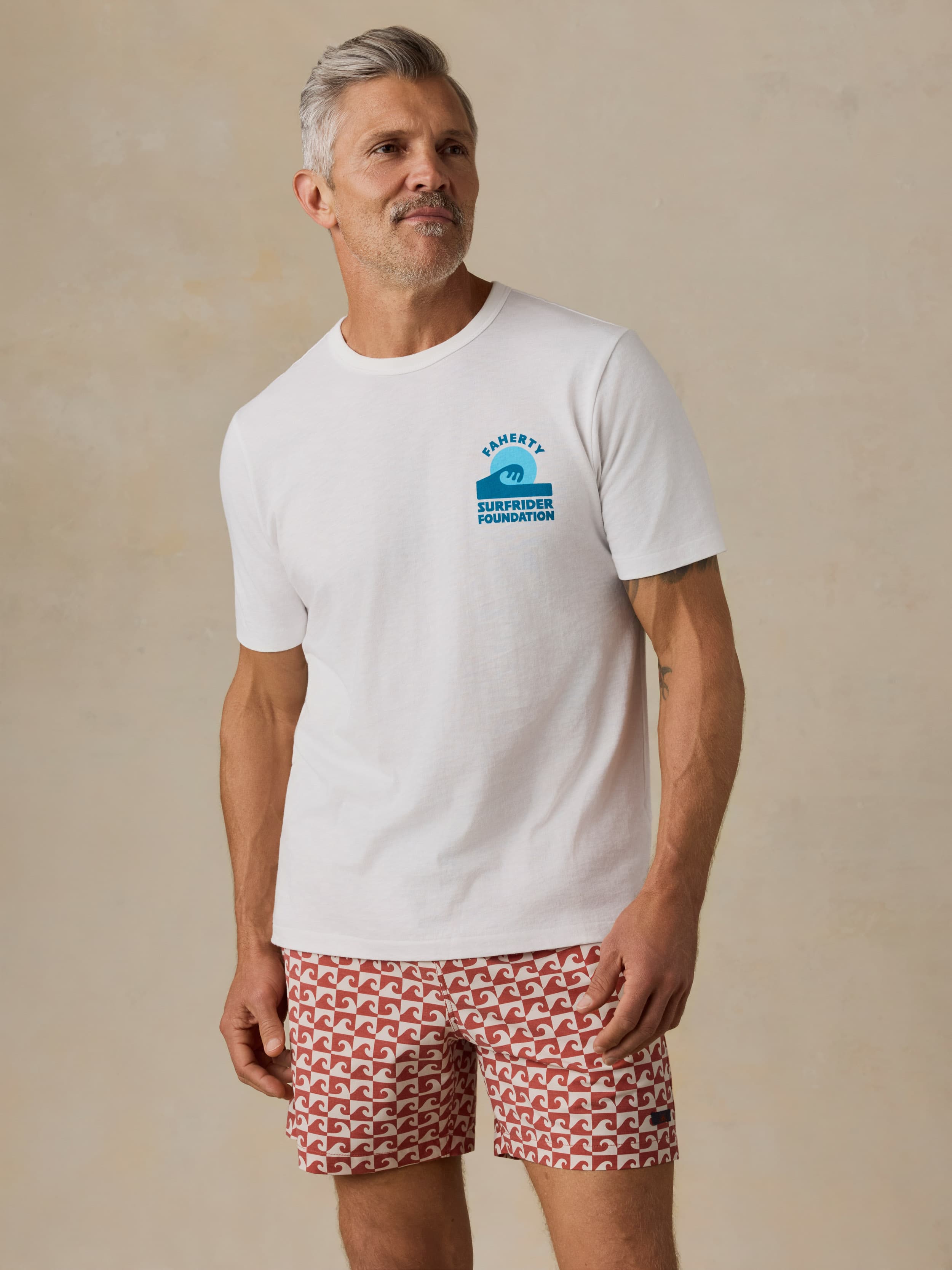Man wearing a white t-shirt with a Faherty x Surfrider Foundation logo and patterned shorts on a beige background