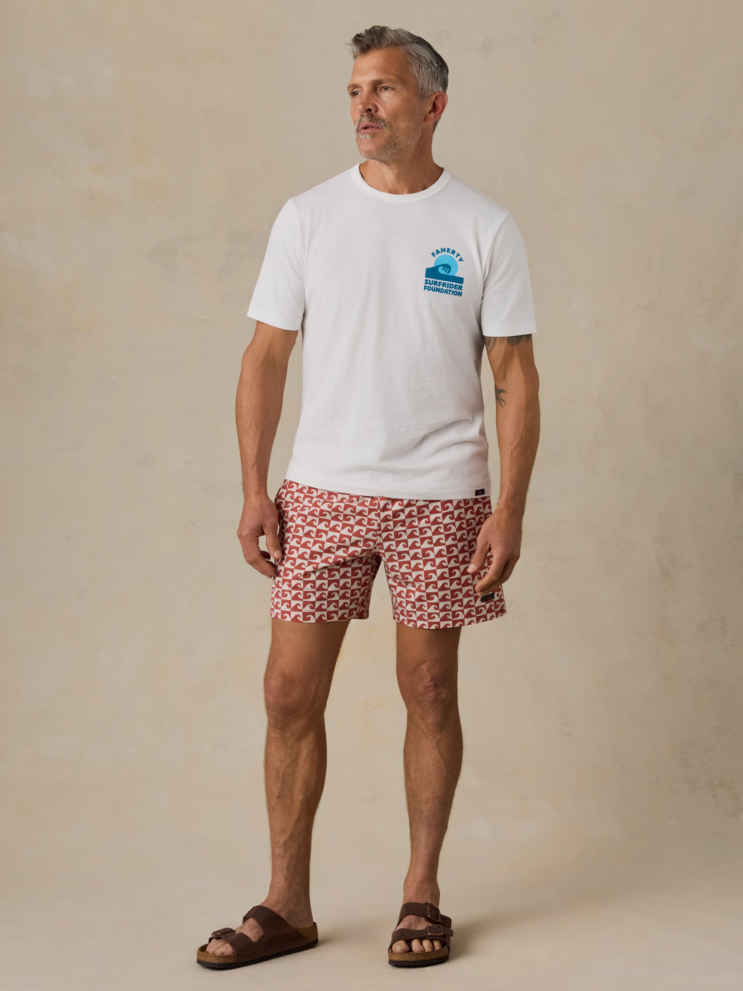 Man wearing a white t-shirt with a Faherty x Surfrider Foundation logo and patterned shorts on a beige background
