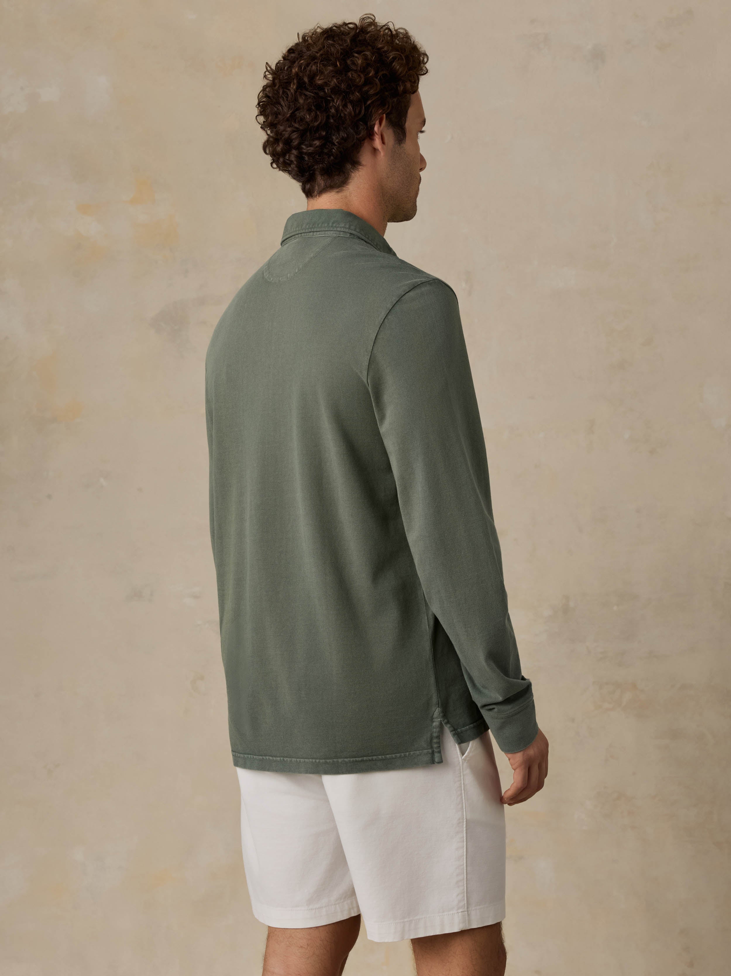 Man wearing a green long-sleeve shirt and white shorts against a beige wall.