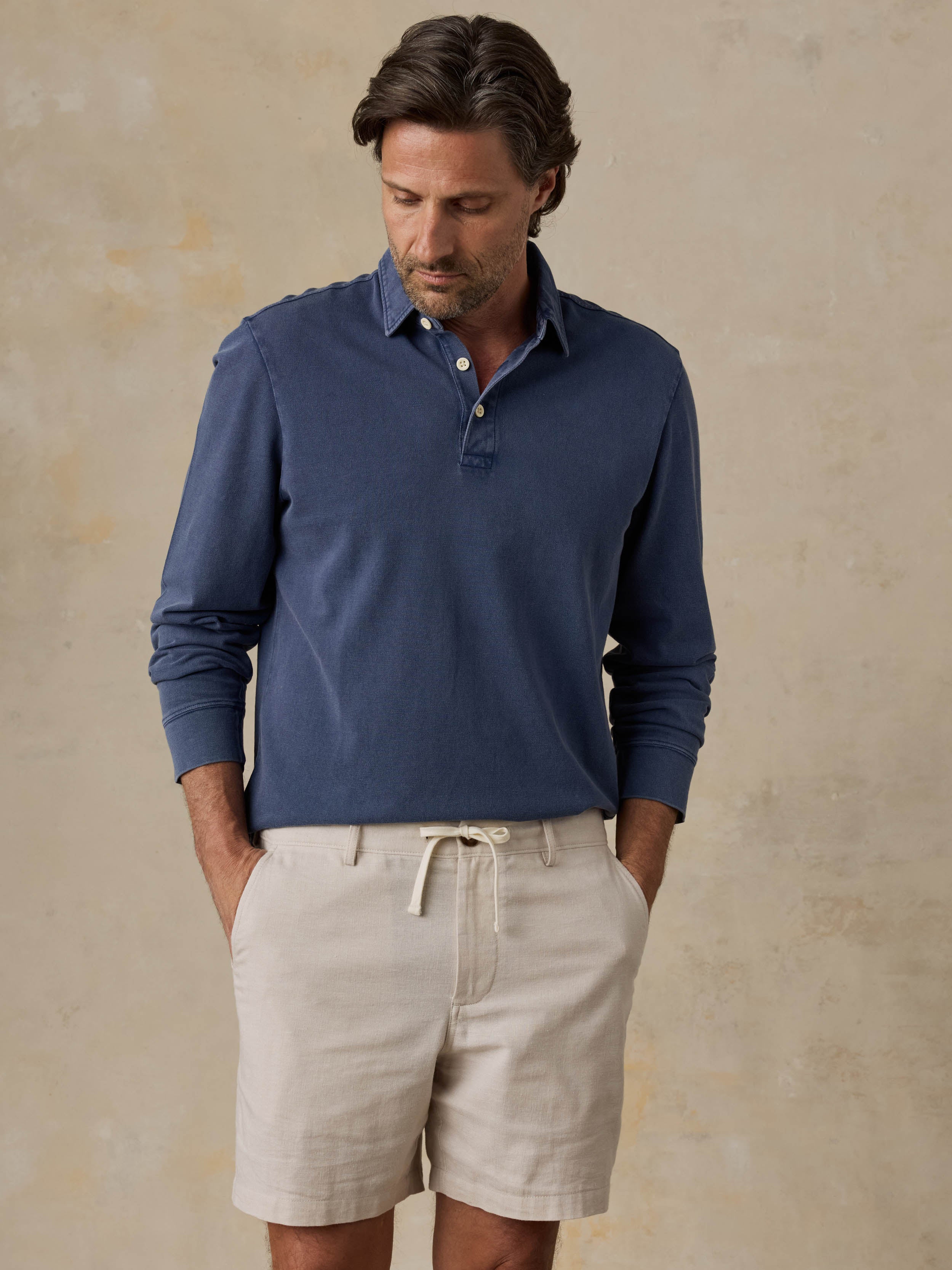 Man wearing a blue long-sleeve polo shirt and beige shorts against a beige background