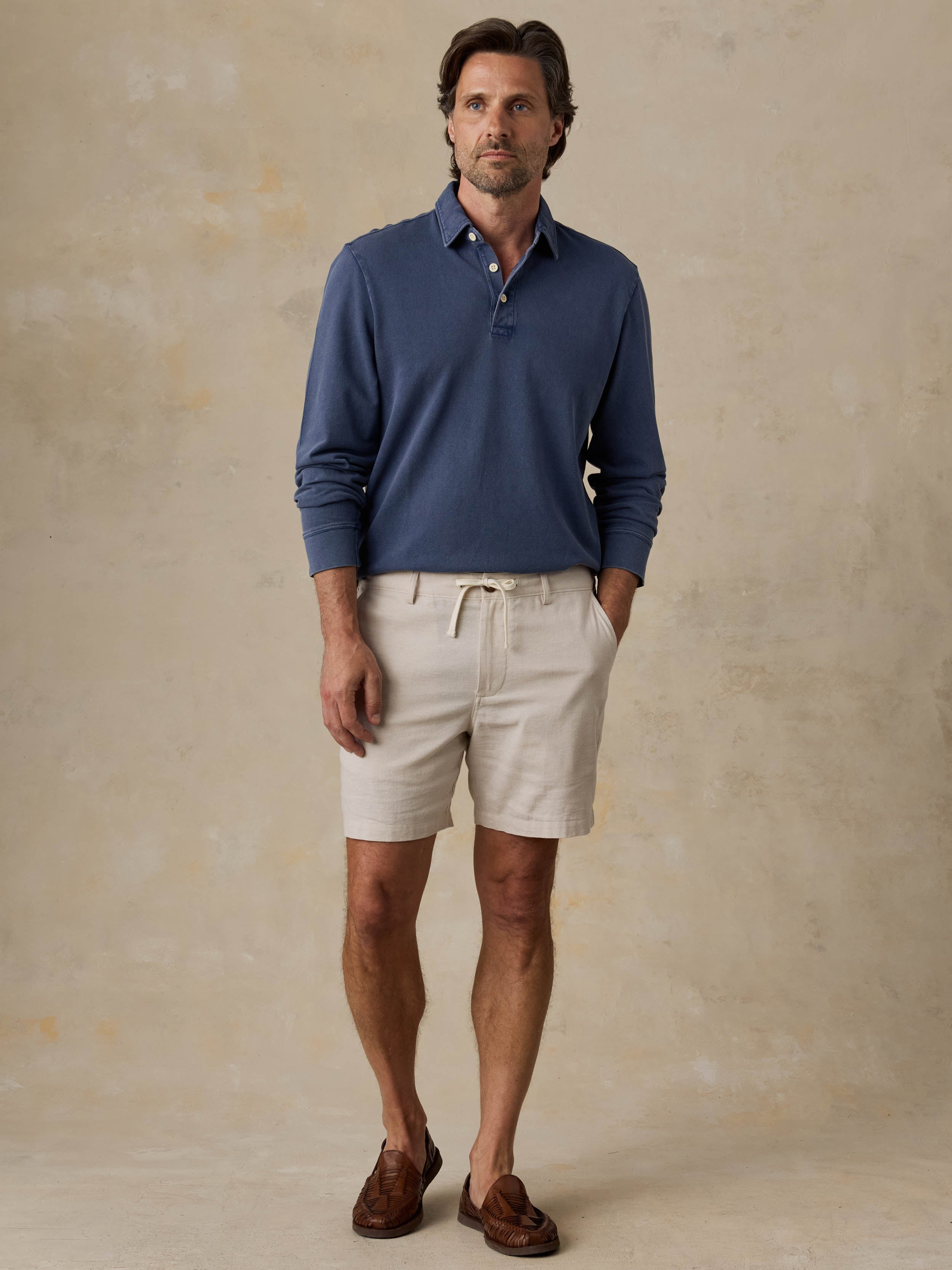 Man wearing a blue long-sleeve polo shirt and beige shorts against a beige background