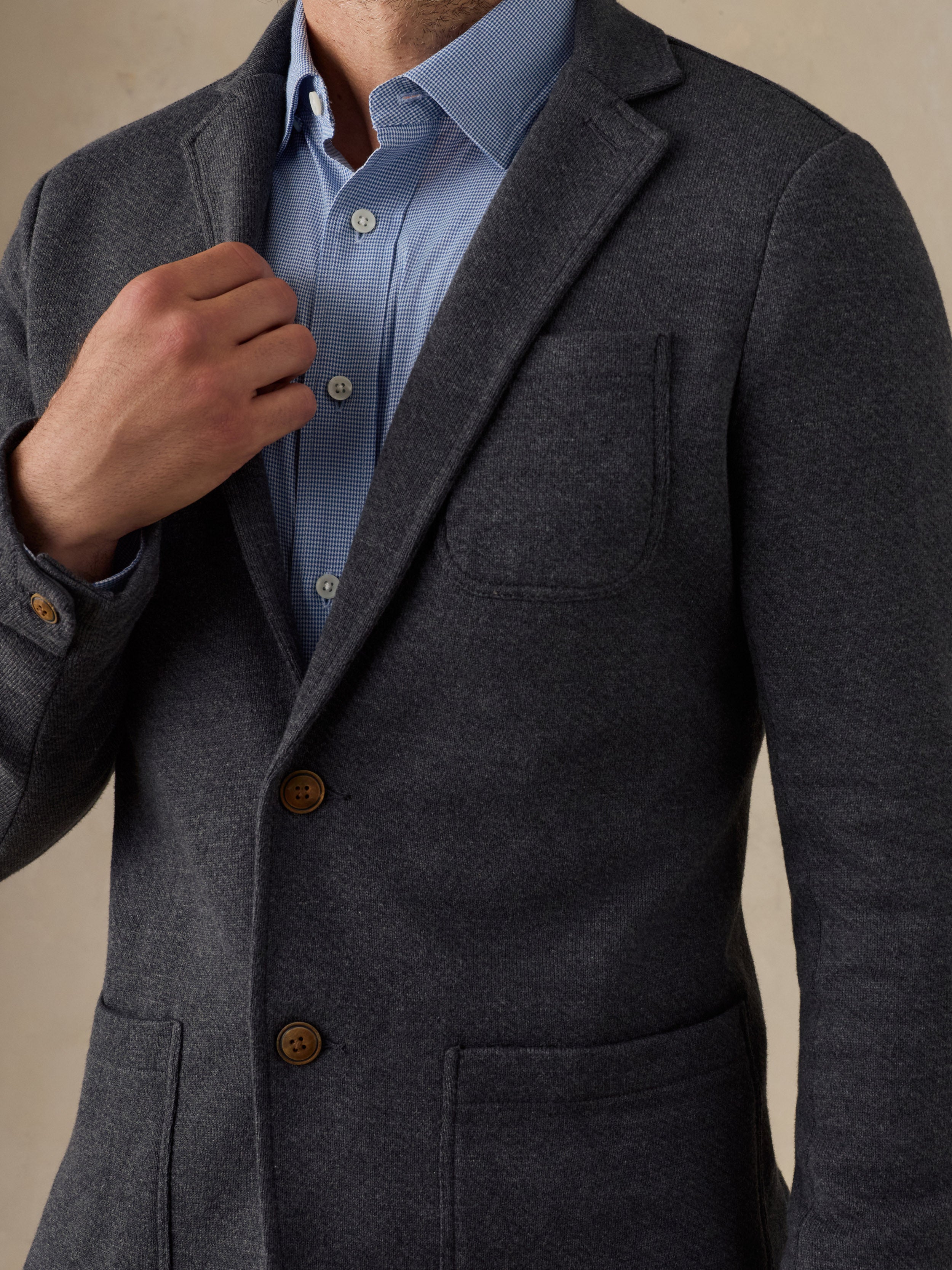 A close-up of a man wearing a dark gray textured blazer over a light blue button-down shirt, with his hand adjusting the collar.
