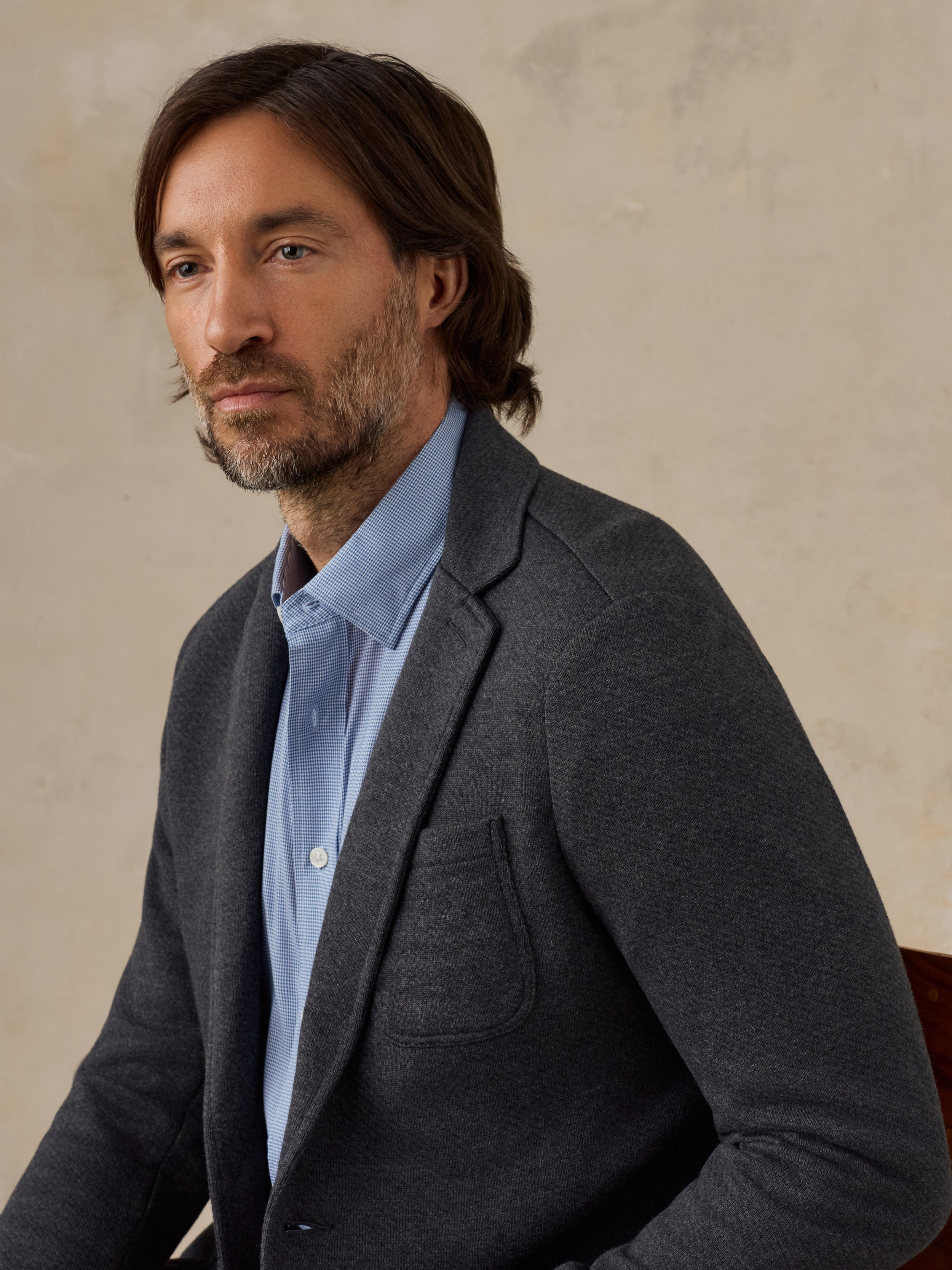 A man wearing a dark gray knit blazer over a light blue button-down shirt, photographed against a textured beige wall.