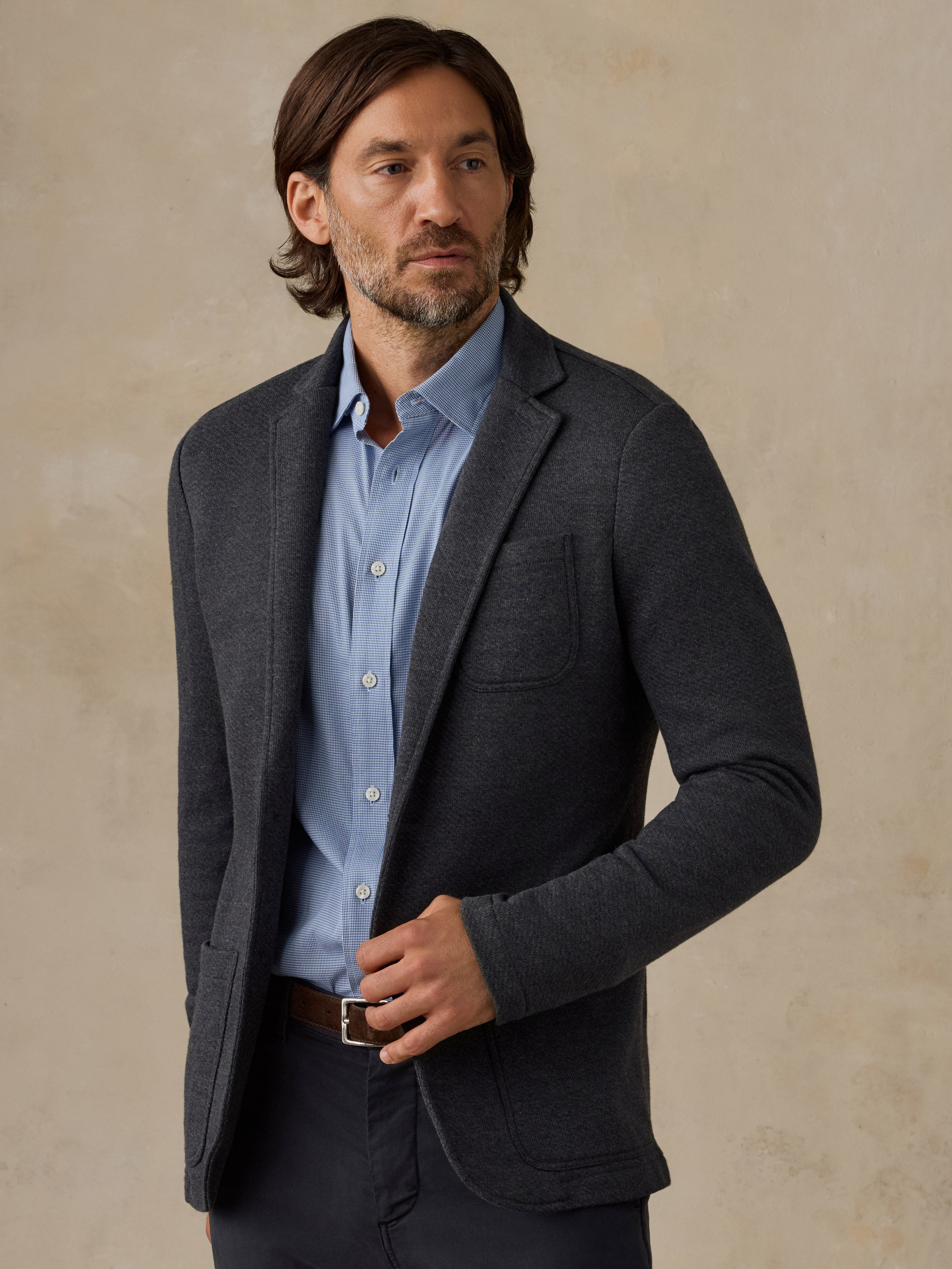 A man wearing a dark charcoal gray textured blazer over a light blue button-down shirt, paired with dark trousers and a brown leather belt, standing against a textured beige background.