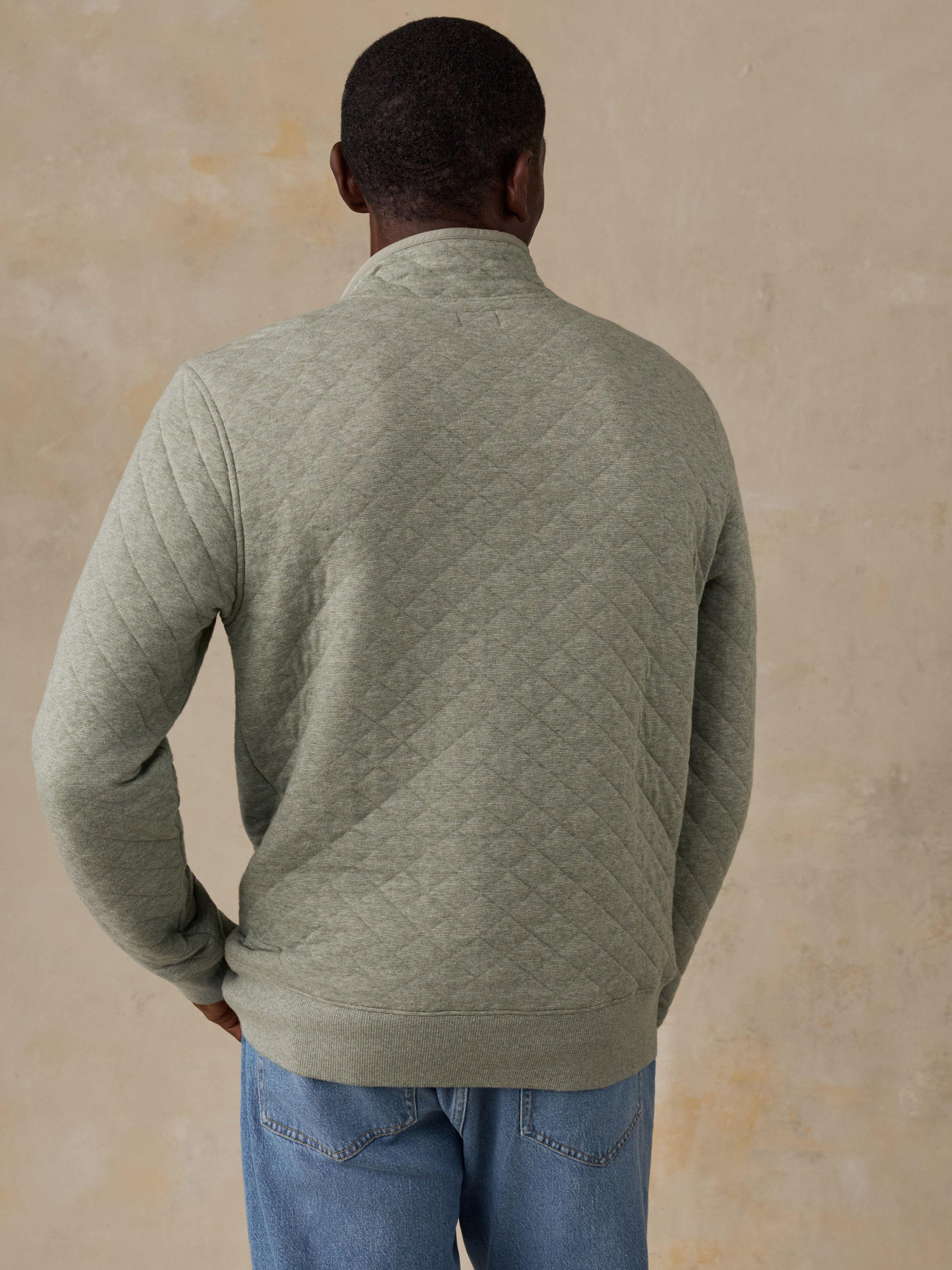 A man wearing a sage green quilted long-sleeve pullover and blue jeans, standing next to a wooden chair against a textured beige wall.