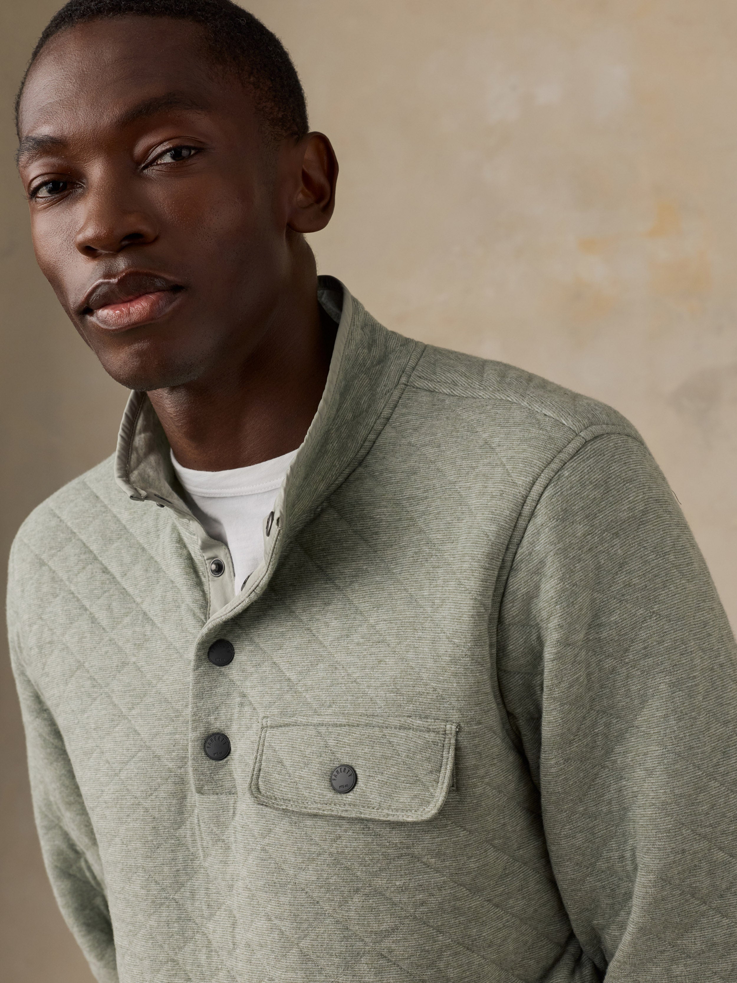 A man wearing a sage green quilted long-sleeve pullover over a white t-shirt, standing next to a wooden chair against a textured beige wall.
