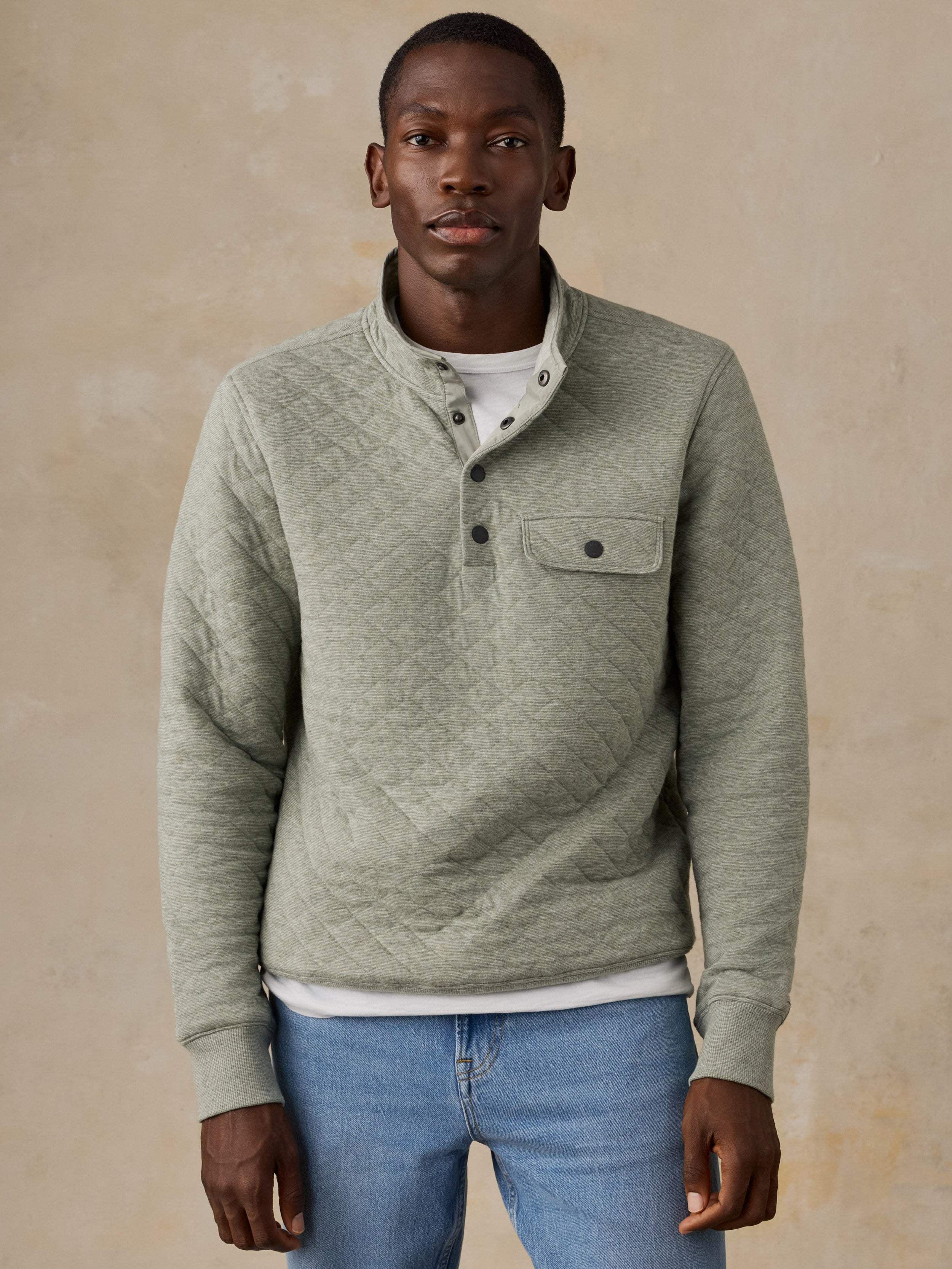 A man wearing a light sage green quilted long-sleeve pullover over a white t-shirt and blue jeans, standing against a textured beige background.