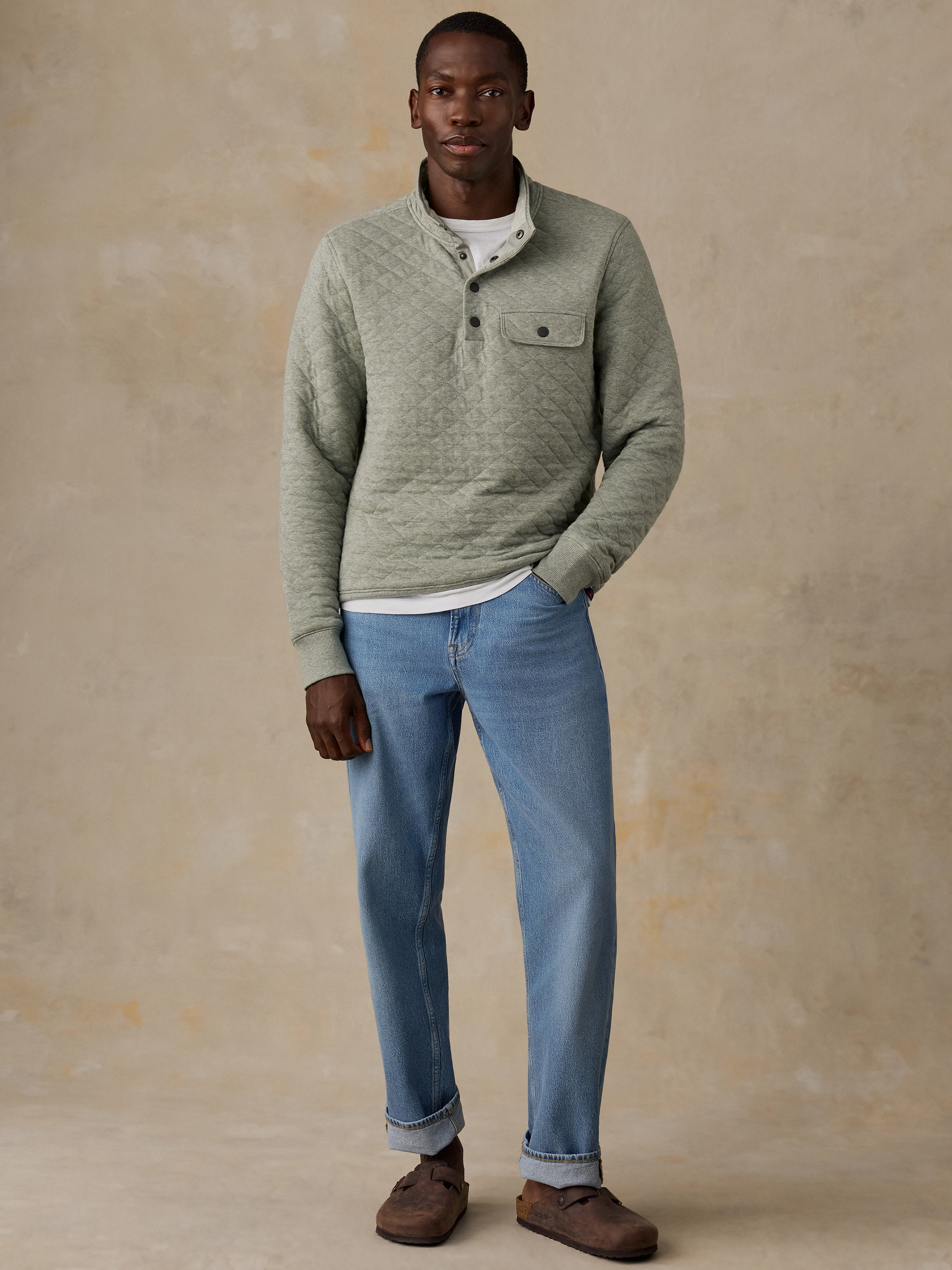 A man wearing a sage green quilted long-sleeve pullover over a white t-shirt and blue jeans, standing next to a wooden chair against a textured beige wall.